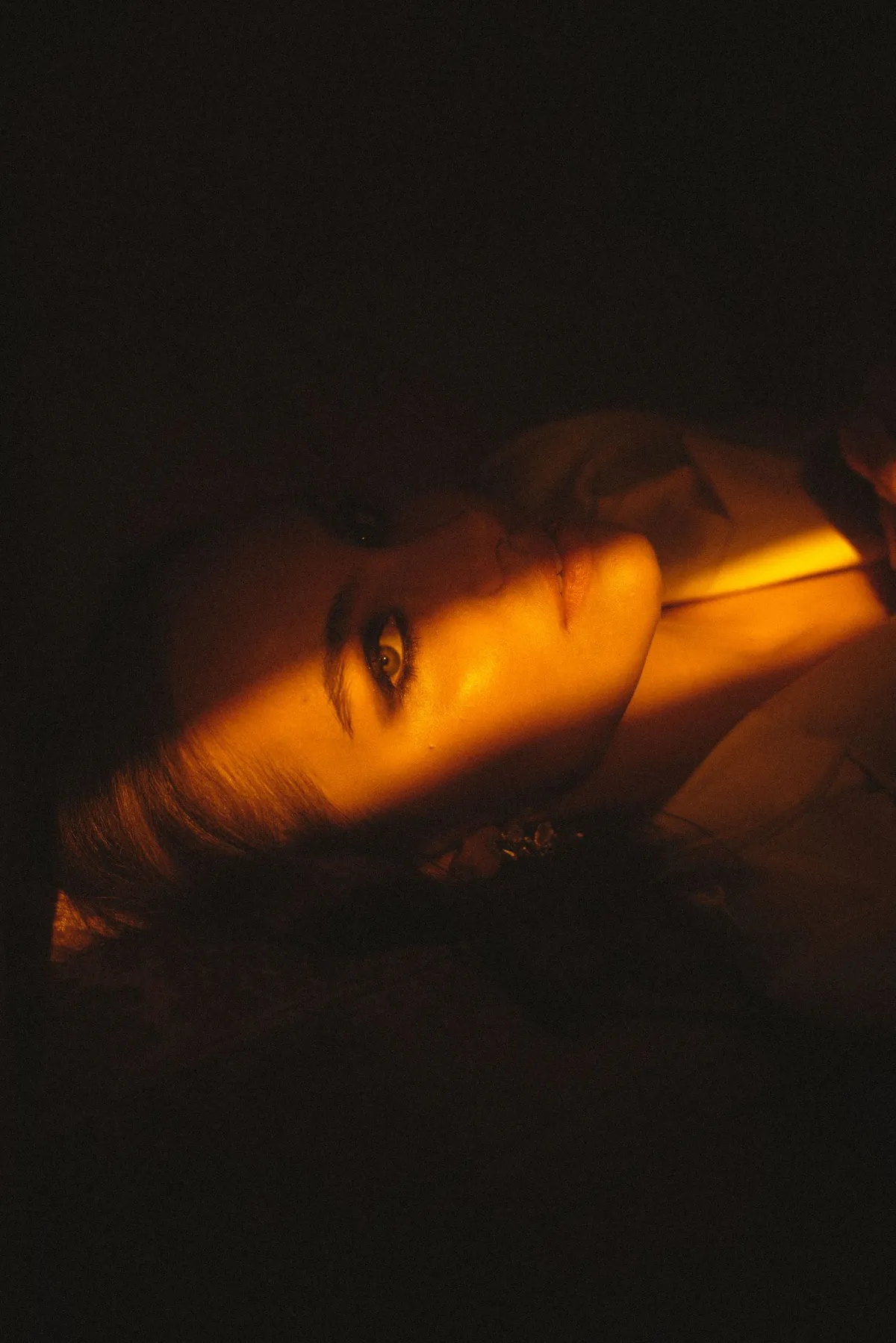 Marie, reclining woman with a strip of orange light across her eyes in darkness, portrait series 2022, photographed by Kilian Amrehn