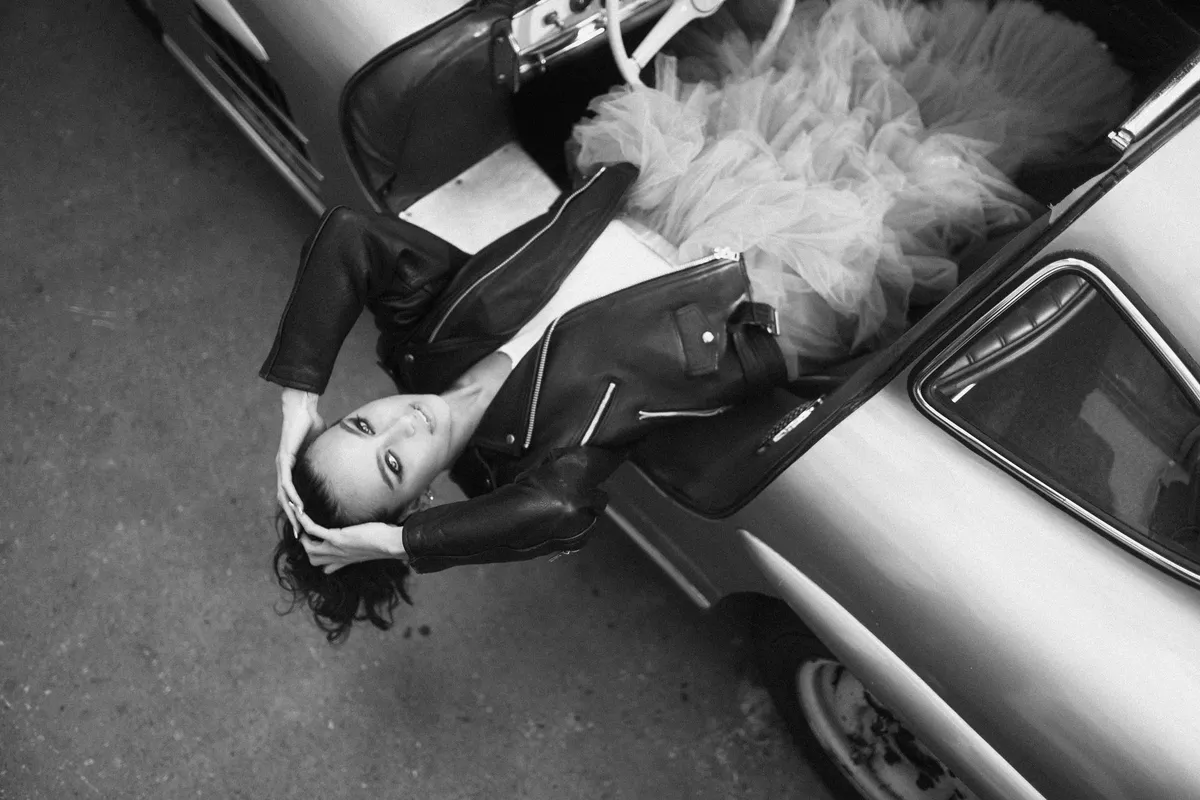 Marie, woman in a leather jacket and tulle skirt lying back out of a vintage car's open door, black and white, portrait series 2022, photographed by Kilian Amrehn