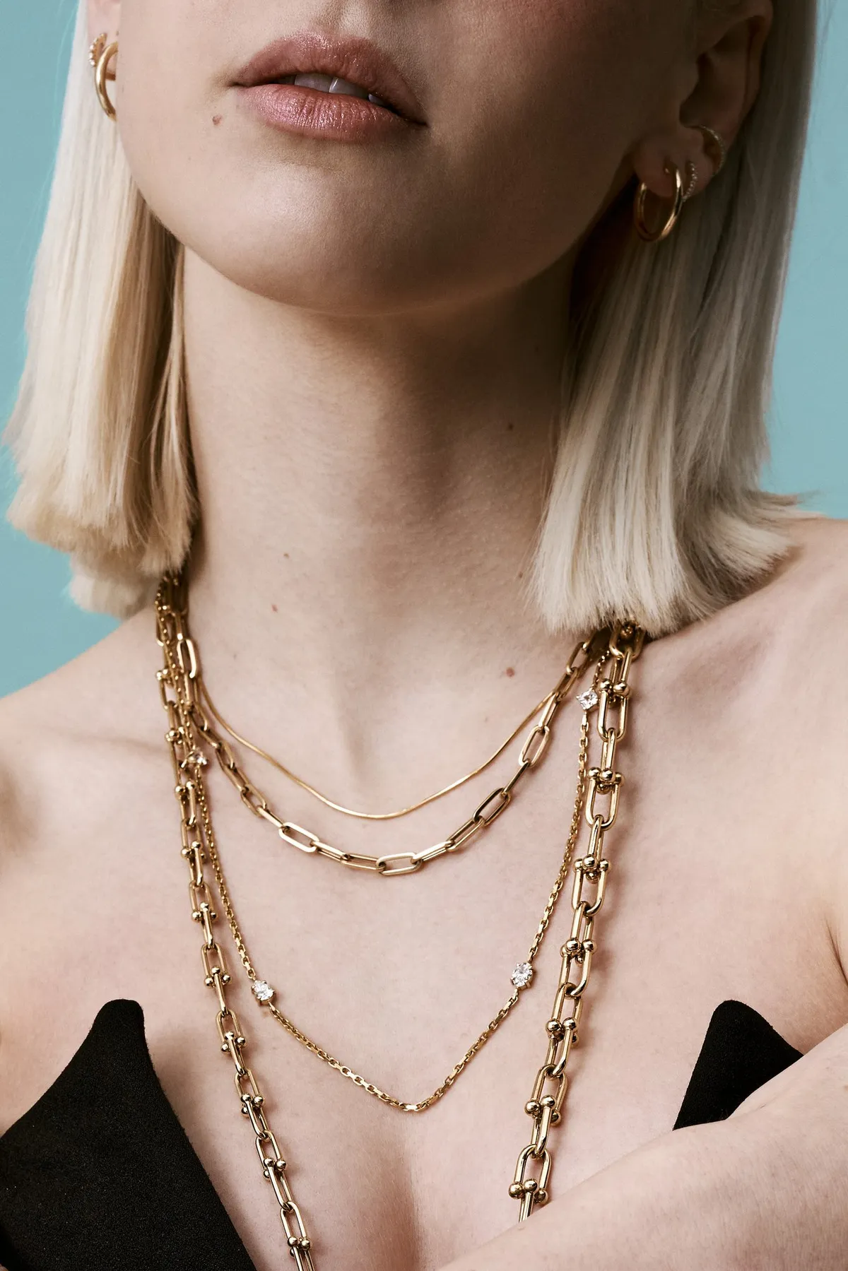 Monano, blonde model wearing layered gold link chains and hoop earrings, black top against pale blue, jewellery campaign 2024, photographed by Kilian Amrehn
