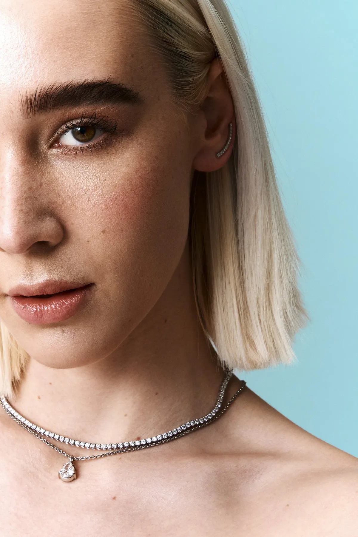 Monano, blonde model wearing a tennis necklace with a pear-cut diamond pendant, pale blue background, jewellery campaign 2024, photographed by Kilian Amrehn