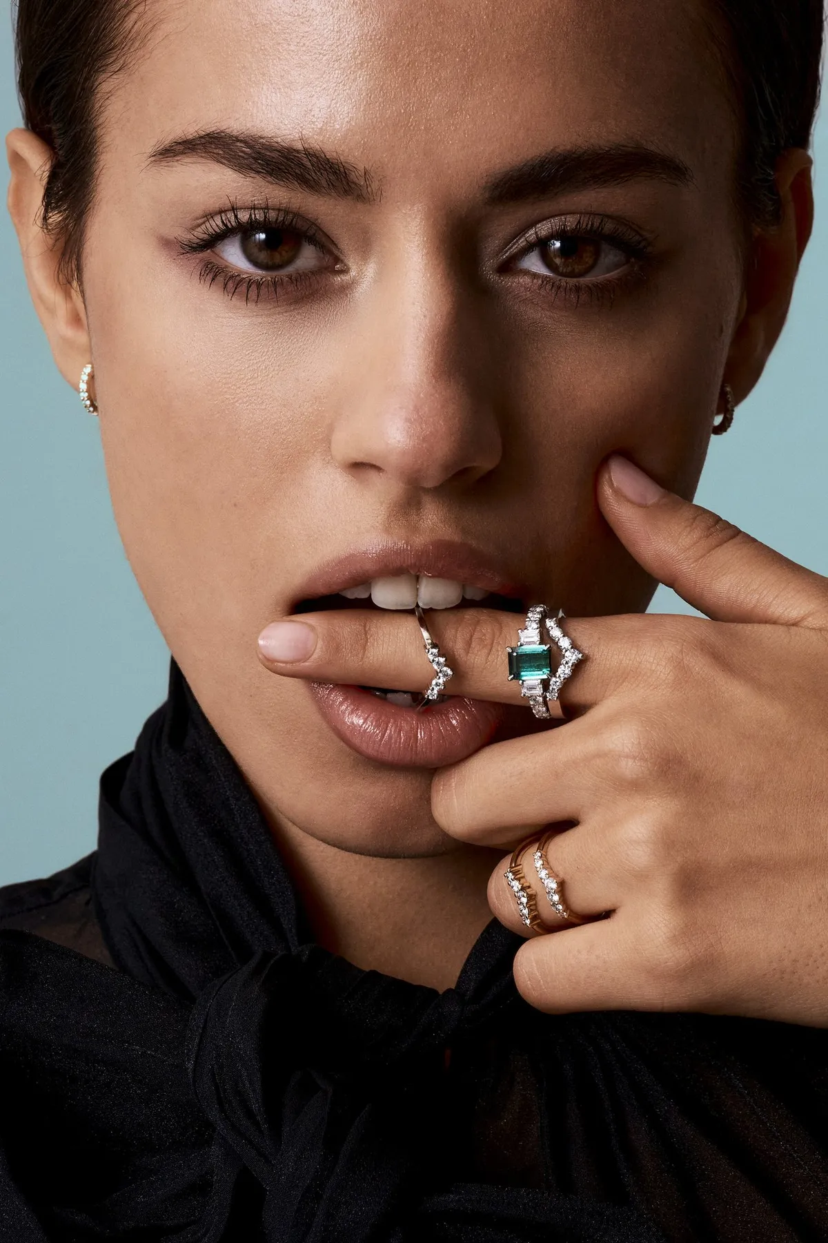 Monano, dark-haired model holding fingers with diamond rings and a green gemstone ring to her lips, jewellery campaign 2024, photographed by Kilian Amrehn