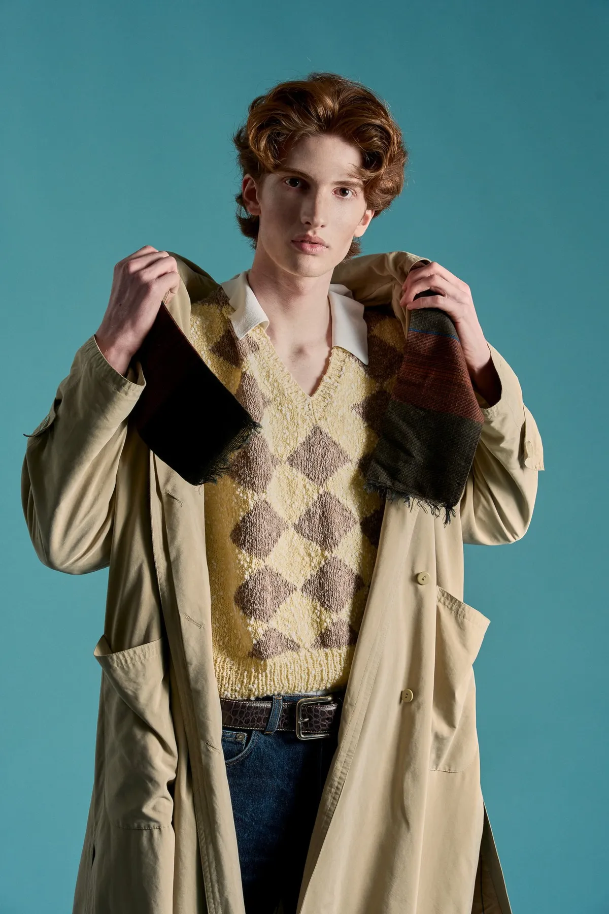 New Icons' Club Magazine, curly-haired model in a trench coat and argyle vest holding a fur-trimmed collar, teal backdrop, editorial 2nd Hand 2024, photographed by Kilian Amrehn