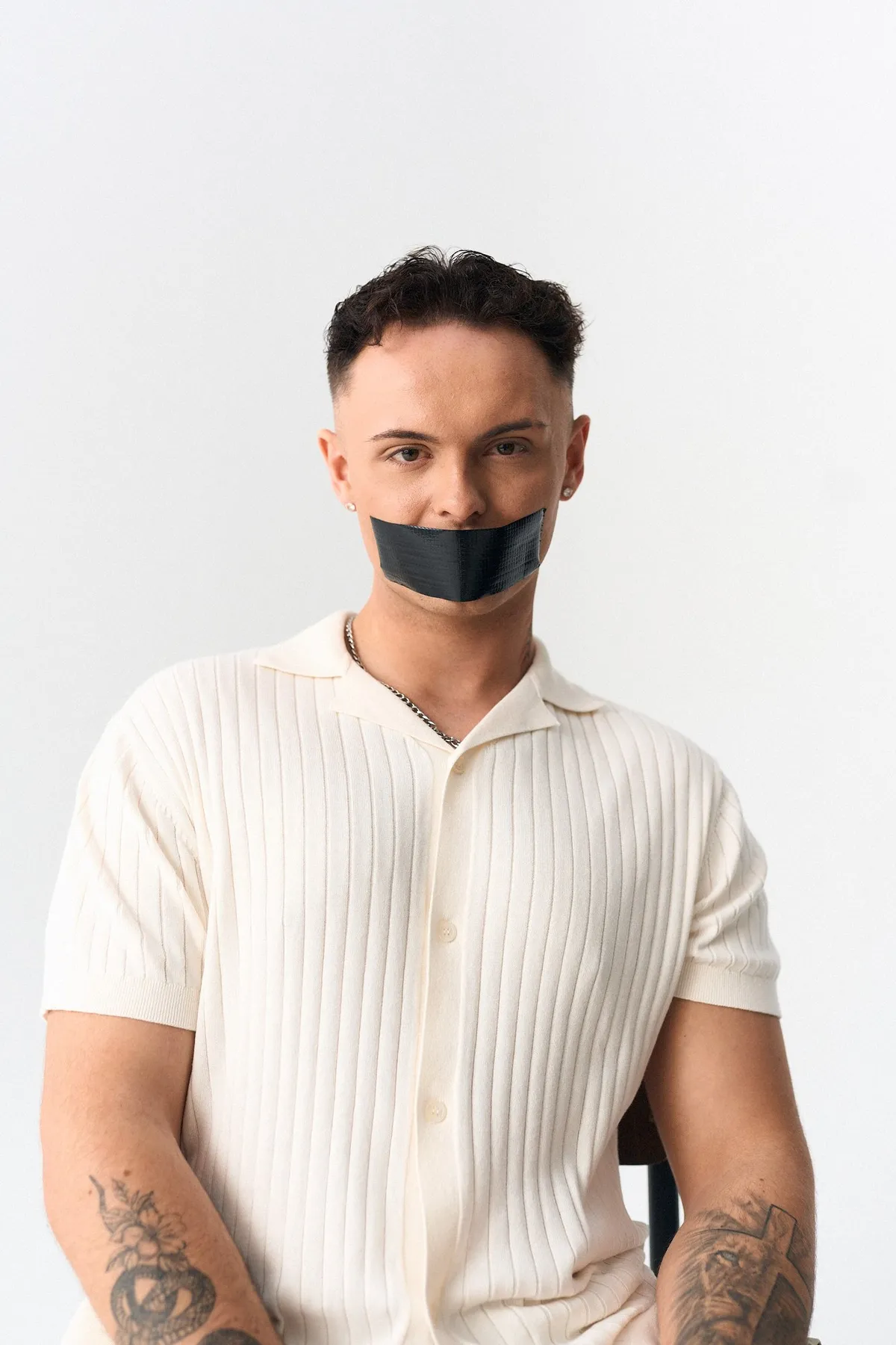 #niewiederleise, tätowierter Mann im cremefarbenen Strickhemd mit Klebeband über dem Mund, frontal vor Weiß, Kampagne 2024, fotografiert von Kilian Amrehn