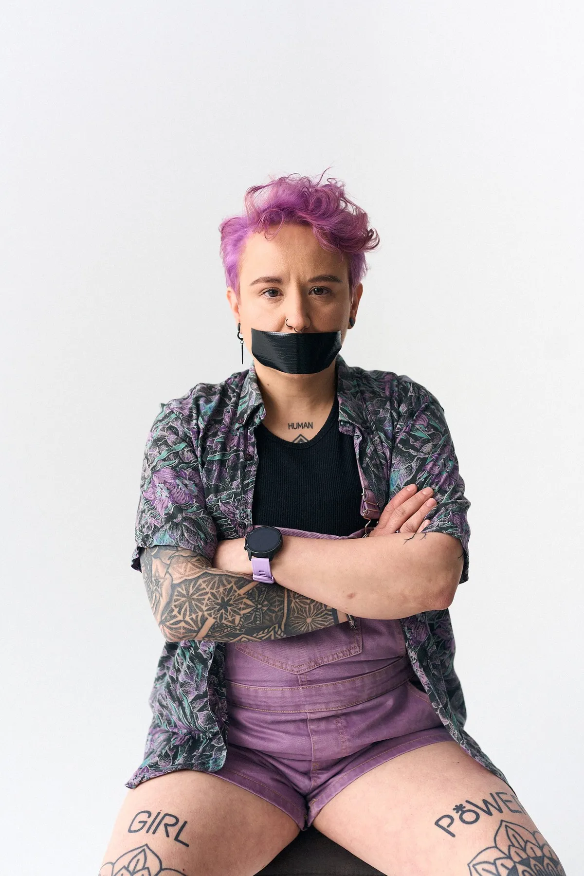 #niewiederleise, Person mit pinkem Haar und verschränkten Armen, Tattoos GIRL und POWER auf den Beinen, Mund abgeklebt, Kampagne 2024, fotografiert von Kilian Amrehn