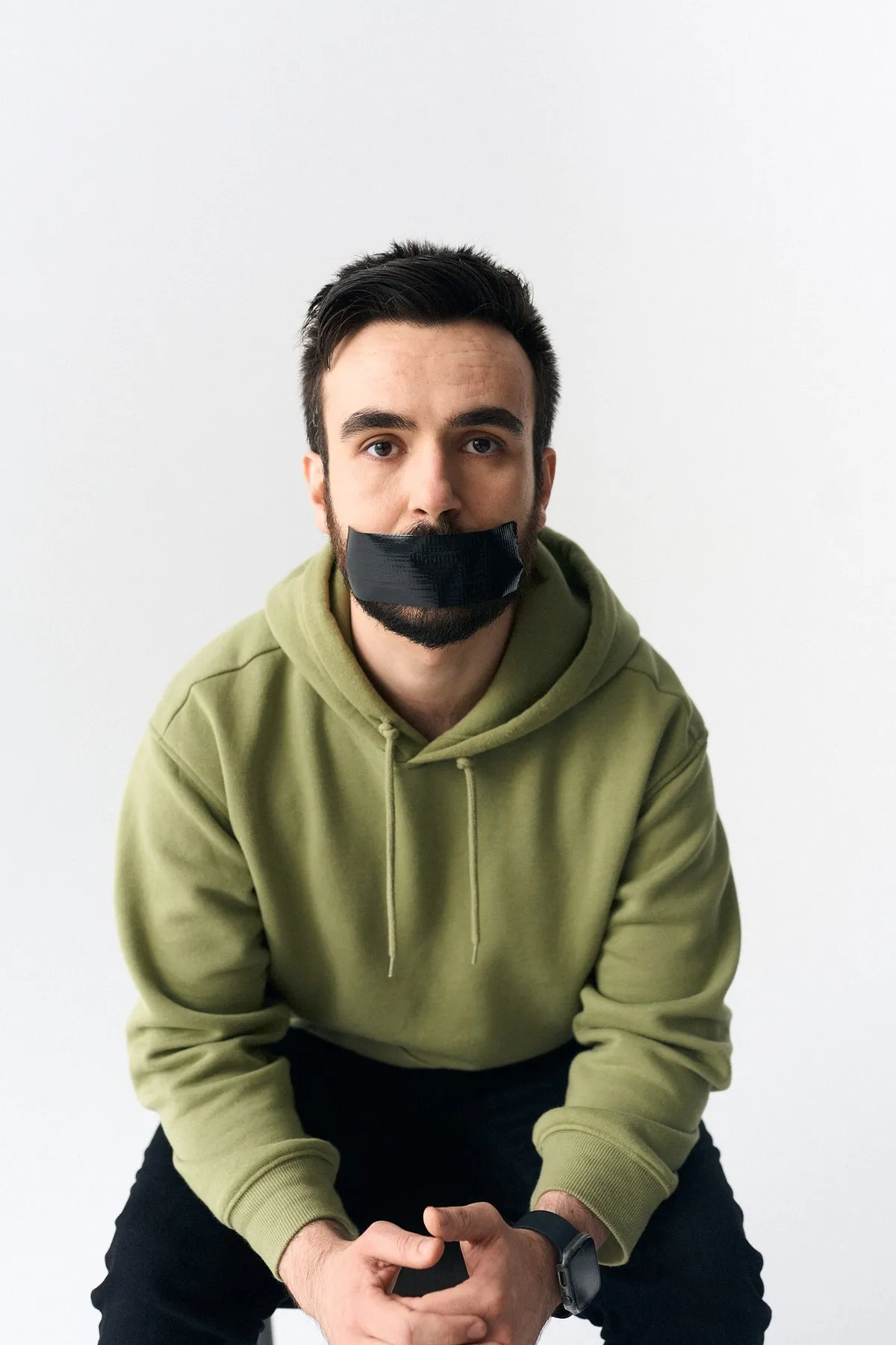 #niewiederleise, Mann im grünen Hoodie sitzt vorgebeugt mit gefalteten Händen, schwarzes Klebeband über dem Mund, Kampagne 2024, fotografiert von Kilian Amrehn