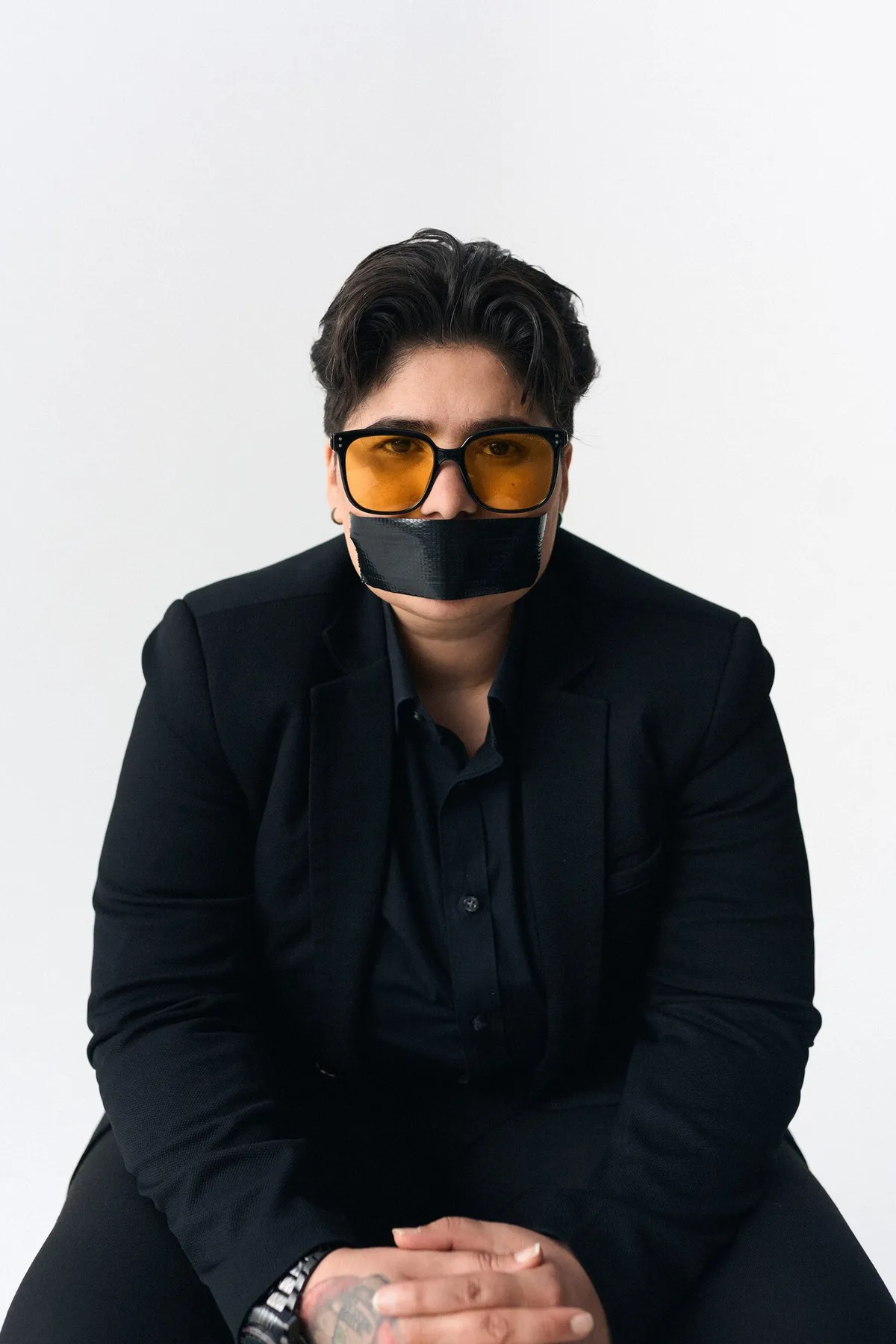 #niewiederleise, Person mit oranger Sonnenbrille im schwarzen Blazer, schwarzes Klebeband über dem Mund, frontal, Kampagne 2024, fotografiert von Kilian Amrehn