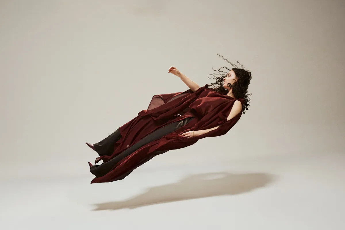 Ozean der Stille, curly-haired model in a burgundy chiffon outfit appearing to float backwards above the floor, bright studio, fashion story 2026, photographed by Kilian Amrehn