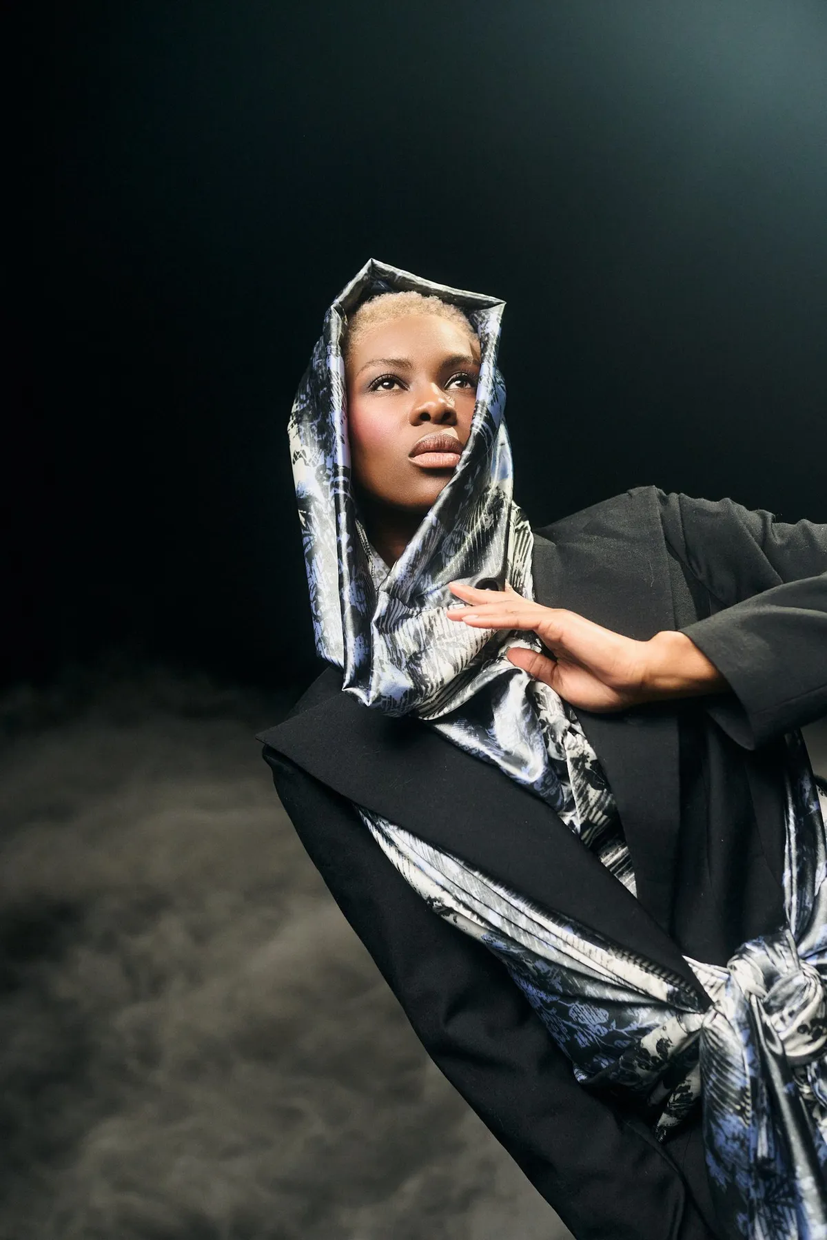Ozean der Stille, Model mit blondem Haar unter glänzender Kapuze aus bedrucktem Satin vor Schwarz, Bodennebel, Modestrecke 2026, fotografiert von Kilian Amrehn