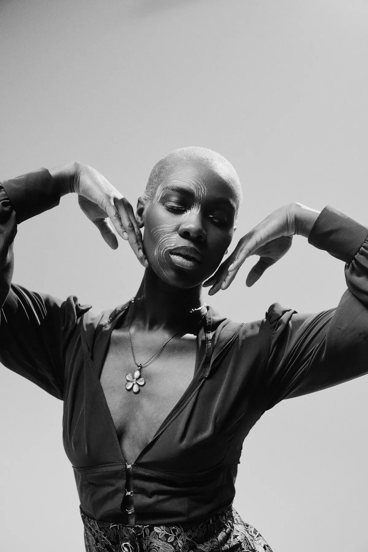 Ozean der Stille, Model mit kurzem hellem Haar und Blütenkette rahmt das Gesicht mit den Händen, Schwarzweiß-Studioporträt, Modestrecke 2026, fotografiert von Kilian Amrehn