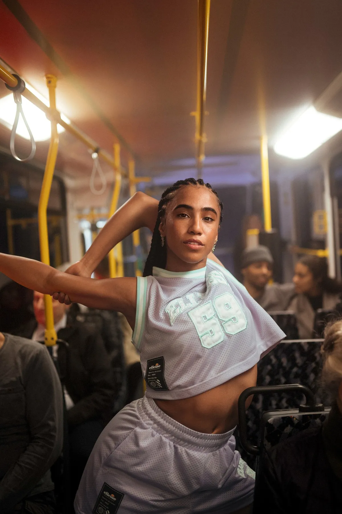 Peek & Cloppenburg x Ellesse, model with long braids posing in a lilac Ellesse crop top and shorts among tram passengers, campaign 2022, photographed by Kilian Amrehn