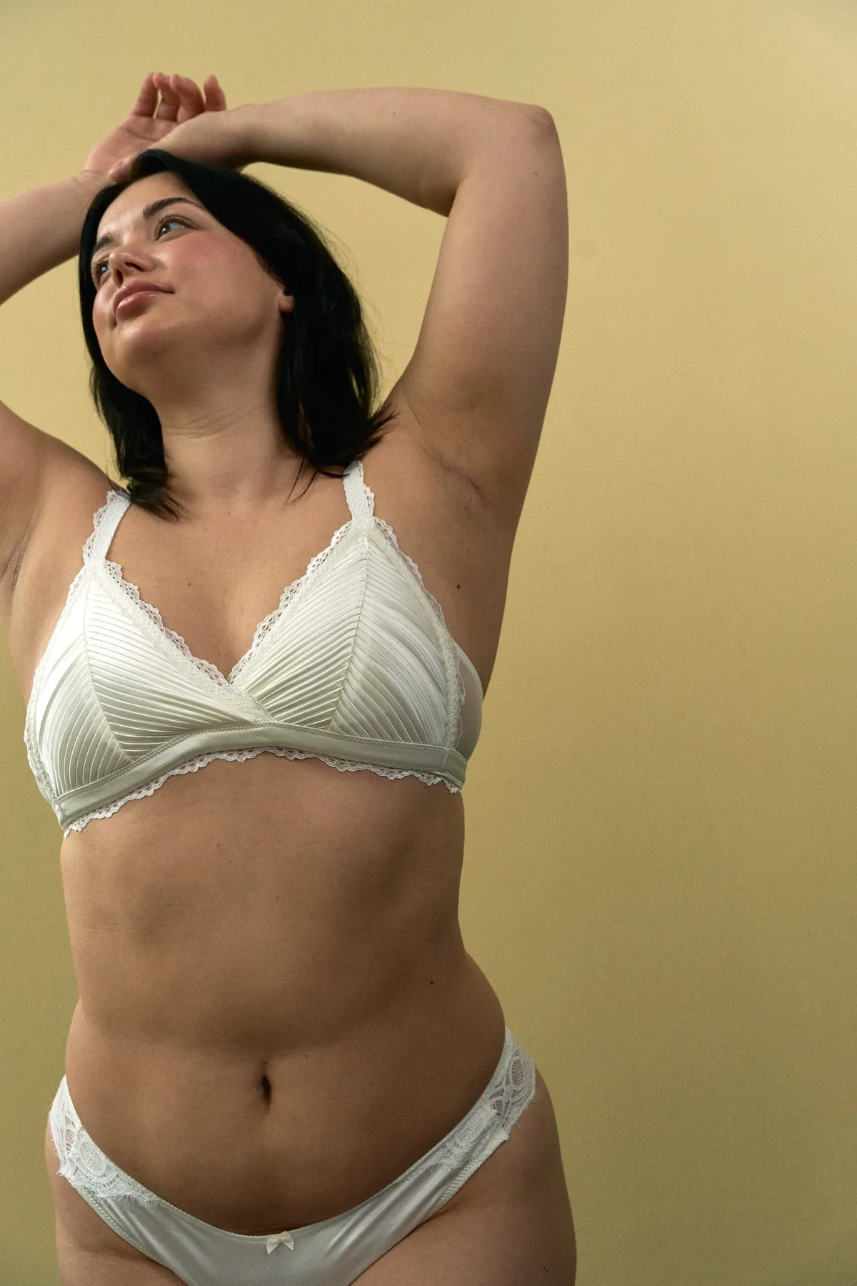 Phiala, model with raised arms in a white pleated bralette and briefs, looking up, yellow background, campaign 2024, photographed by Kilian Amrehn