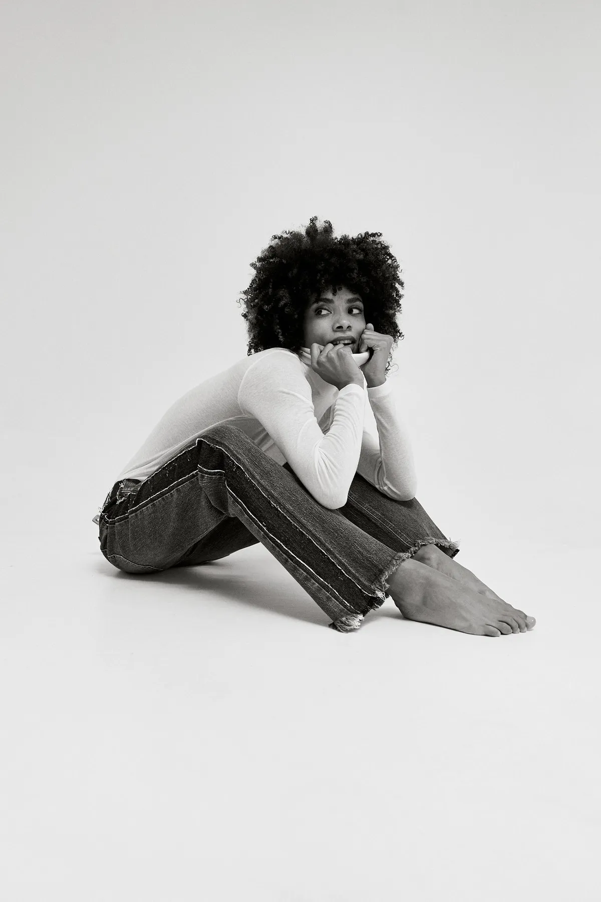PNTS, model with an afro in jeans and a white long sleeve sitting barefoot on the floor, chin on hands, black and white, campaign 2022, photographed by Kilian Amrehn