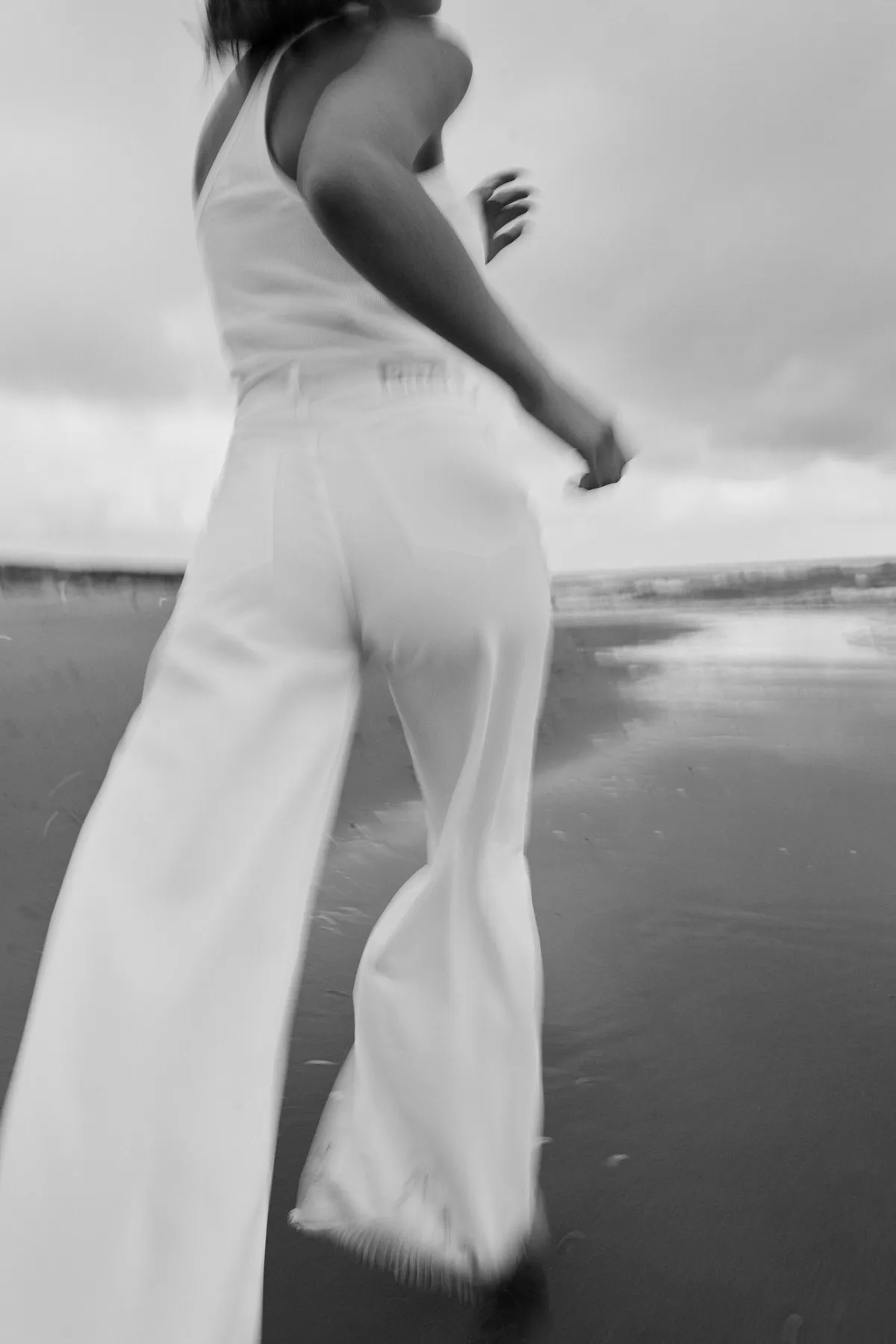 PNTS, model in white wide-leg jeans and top running on the beach, motion blur in black and white, Noordwijk 2023, photographed by Kilian Amrehn