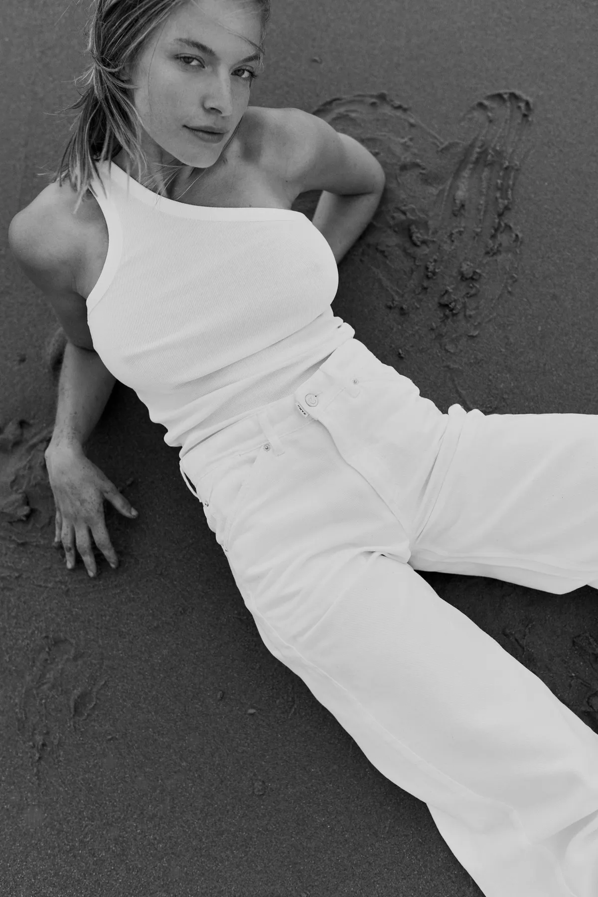 PNTS, model in a white one-shoulder top and white jeans lying in the sand, looking at the camera, black and white, Noordwijk 2023, photographed by Kilian Amrehn
