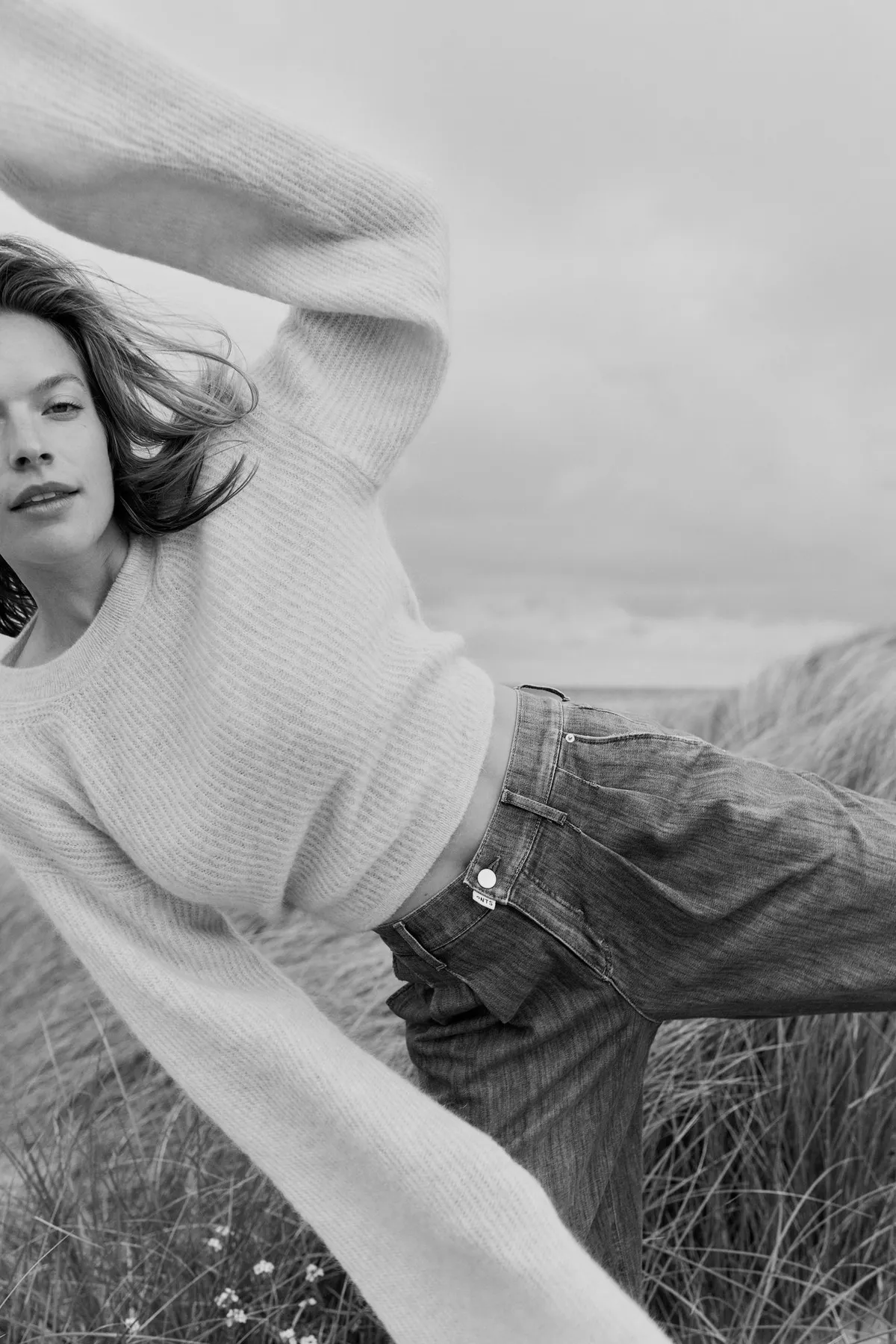 PNTS, model in a light knit sweater and jeans stretching in the dunes, hair in the wind, black and white, Noordwijk 2023, photographed by Kilian Amrehn