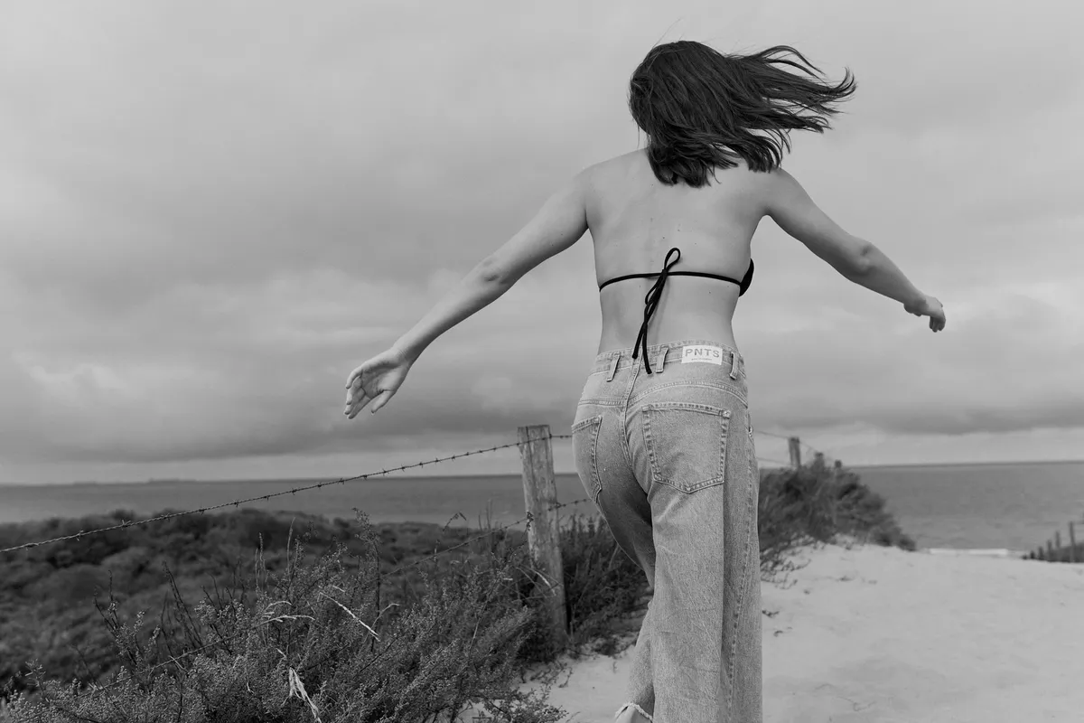 PNTS, model in a bikini top and wide jeans spreading her arms in the dunes by the sea, back view in black and white, Noordwijk 2023, photographed by Kilian Amrehn