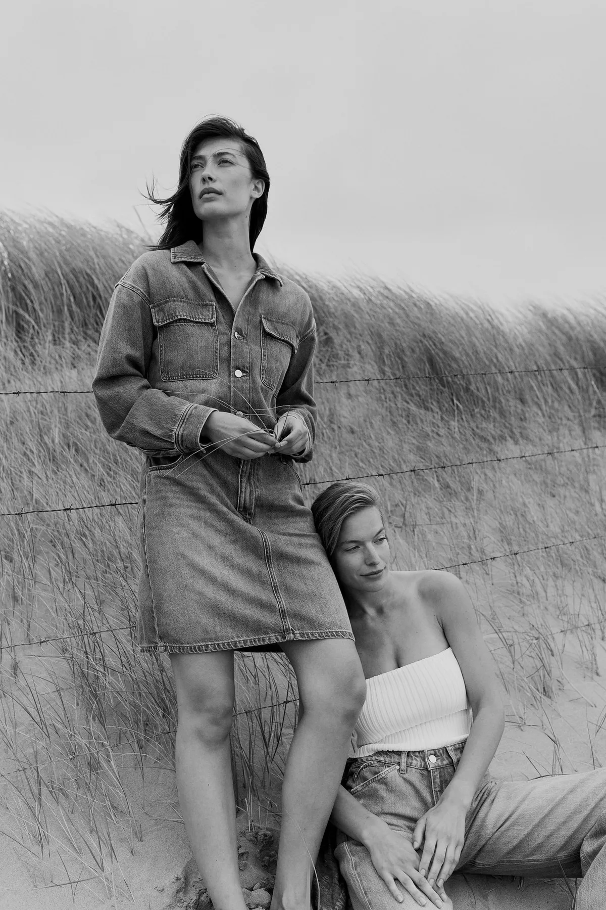 PNTS, model in a denim shirt dress standing in dune grass with a second model in jeans leaning on her, black and white, Noordwijk 2023, photographed by Kilian Amrehn