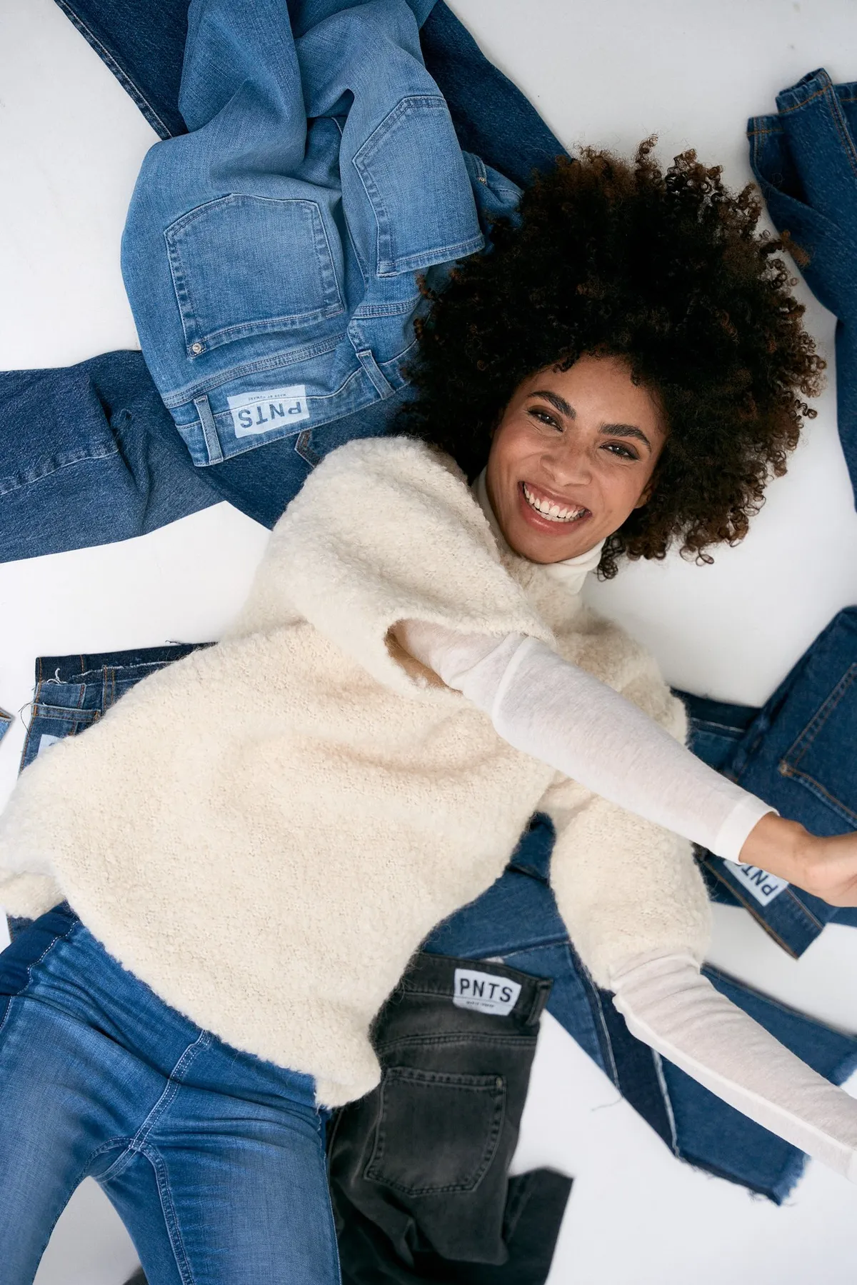 PNTS, laughing model with an afro in a fluffy knit vest lying on spread-out jeans with PNTS patches, campaign 2022, photographed by Kilian Amrehn