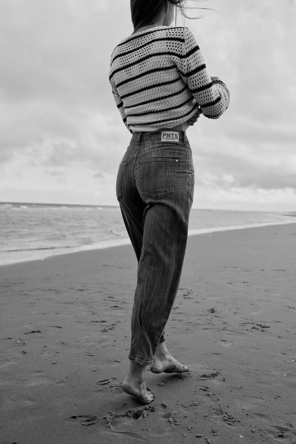 PNTS, model in a striped knit sweater and dark jeans walking barefoot on the beach, black and white, Noordwijk 2023, photographed by Kilian Amrehn