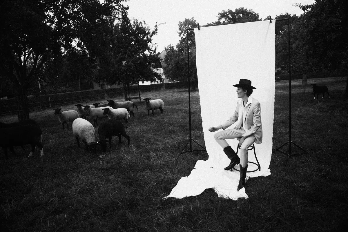 Rock Fairyland, model in a hat and light suit on a stool before a white sheet, surrounded by grazing sheep, black and white, editorial 2021, photographed by Kilian Amrehn