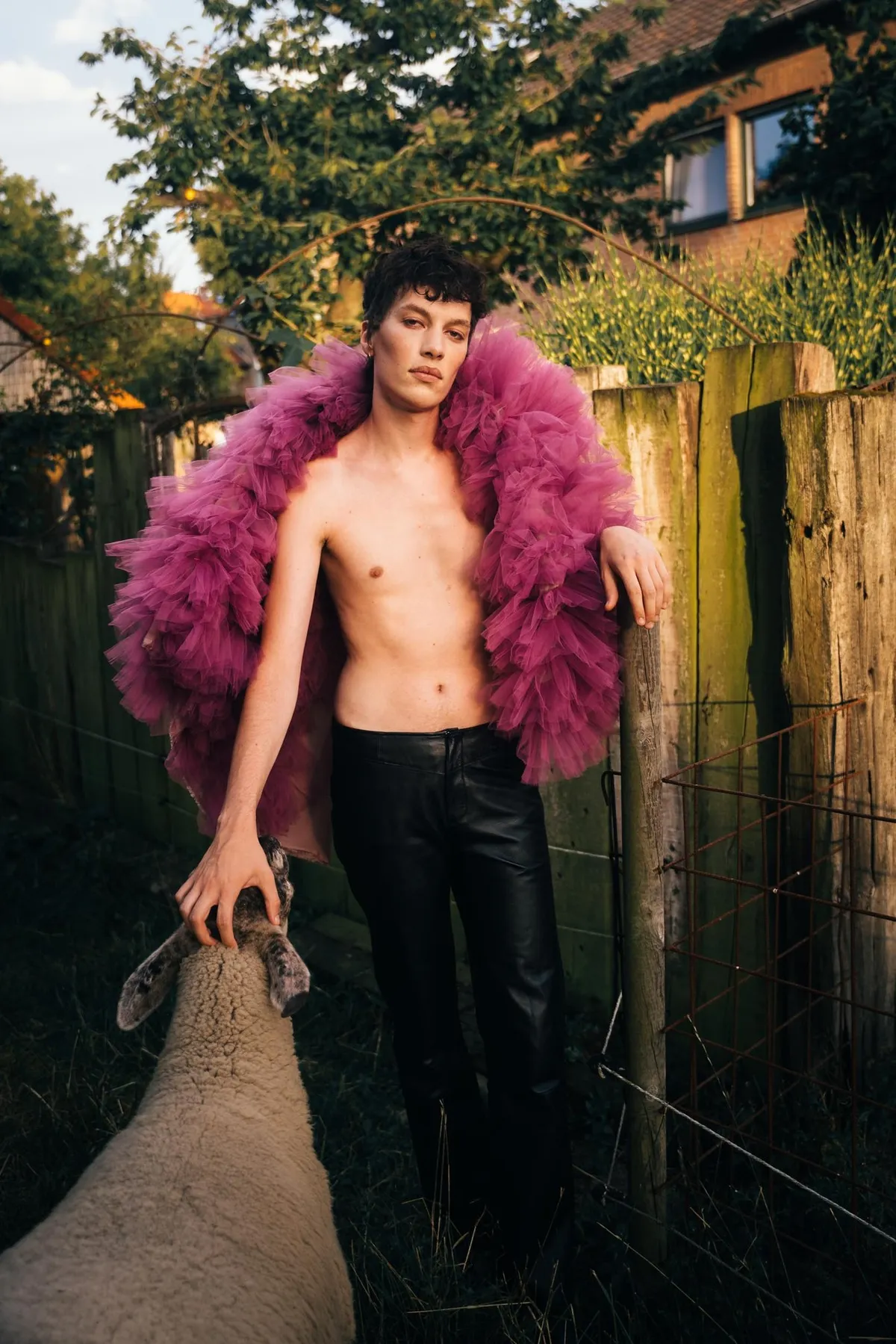 Rock Fairyland, shirtless model in a pink feathered coat and black leather trousers by a wooden fence next to a sheep, editorial 2021, photographed by Kilian Amrehn