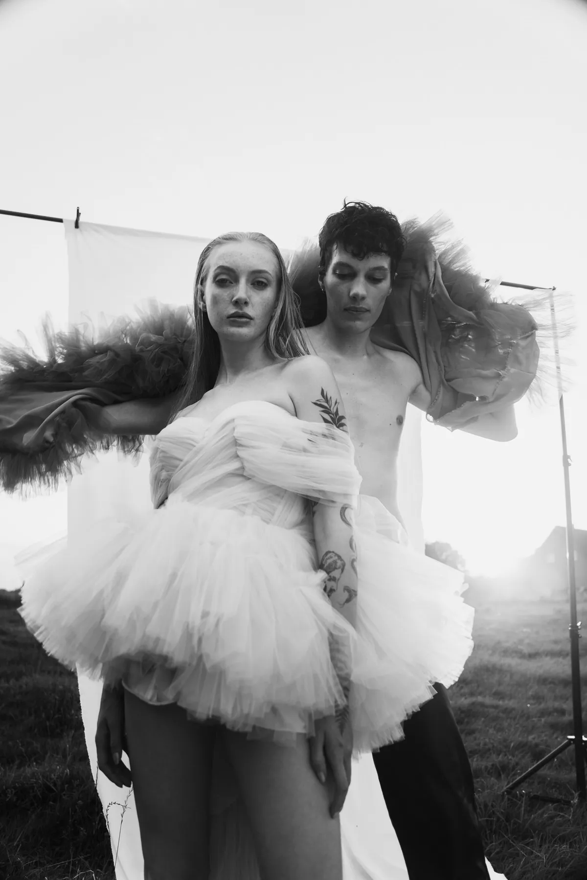 Rock Fairyland, model in a white tulle mini dress in front of a model in a dark tulle cape, backlit meadow, black and white, editorial 2021, photographed by Kilian Amrehn