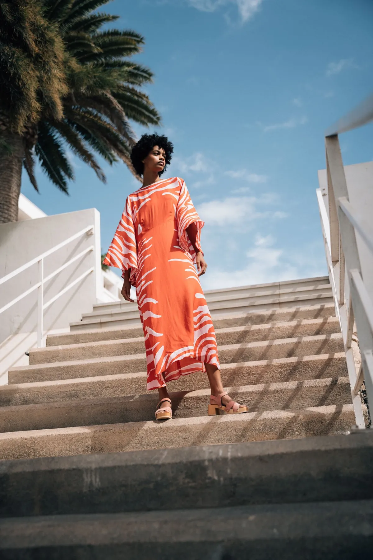 Scholl iconic, model with an afro in an orange and white kaftan and sandals on white stairs under palm trees, campaign Fuerteventura 2021, photographed by Kilian Amrehn