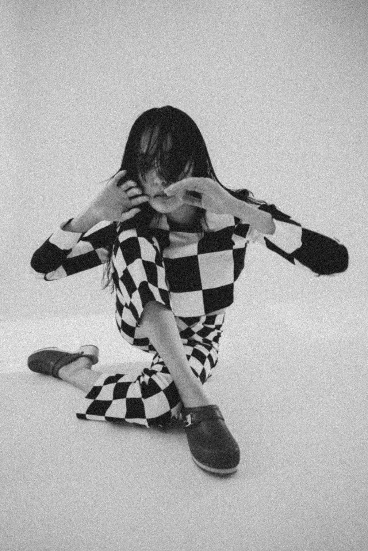 Scholl iconic, model with bangs in a checkerboard outfit and dark clogs kneeling on the floor, grainy black and white, campaign October 2021, photographed by Kilian Amrehn