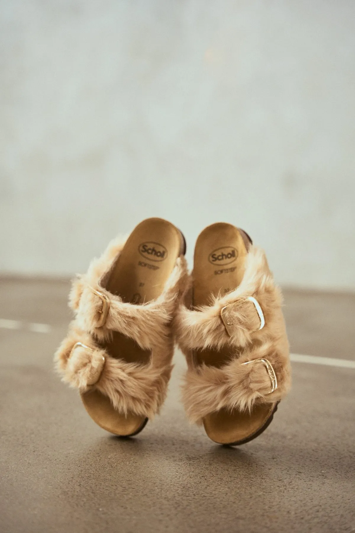 Scholl Comfort, fluffy beige slides with fur straps and cork footbed on a concrete floor, product shot, studio July 2022, photographed by Kilian Amrehn