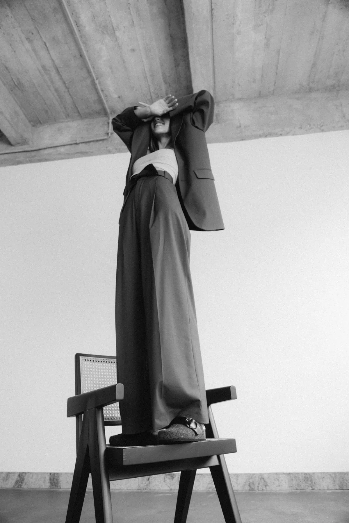 Scholl Comfort, model in a wide dark suit standing on a wooden chair, arm across the face, black and white, studio July 2022, photographed by Kilian Amrehn