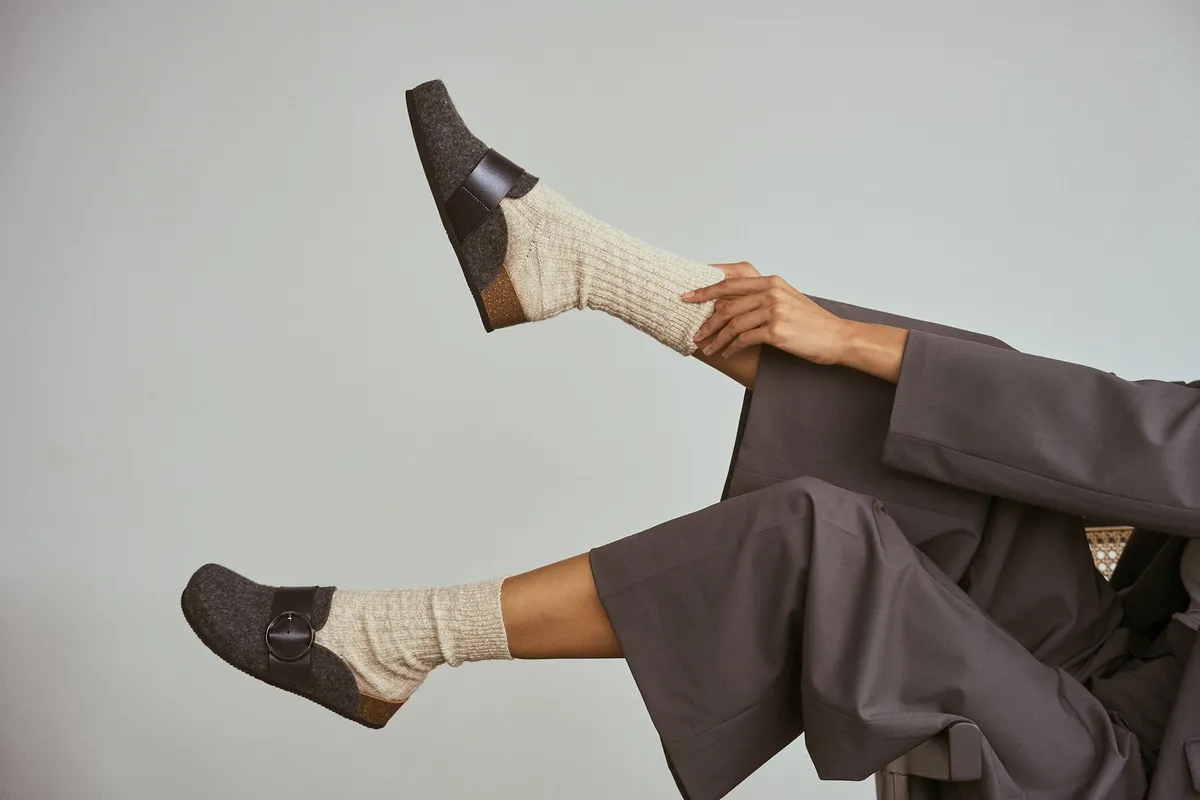 Scholl Comfort, legs in wide grey trousers and knit socks with dark felt clogs raised in the air, light background, studio July 2022, photographed by Kilian Amrehn