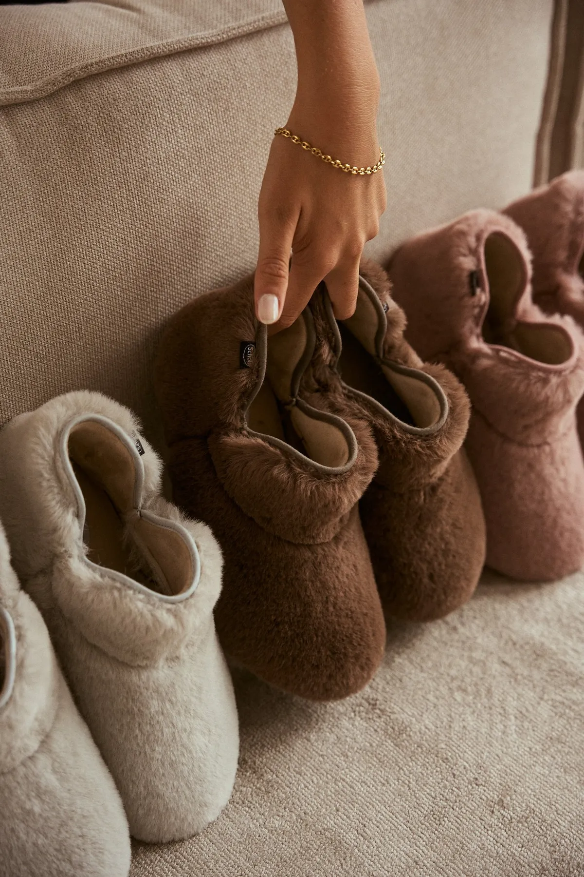 Scholl Comfort, hand with a gold bracelet reaching for fur slippers in cream, brown and rose on a sofa, studio July 2022, photographed by Kilian Amrehn