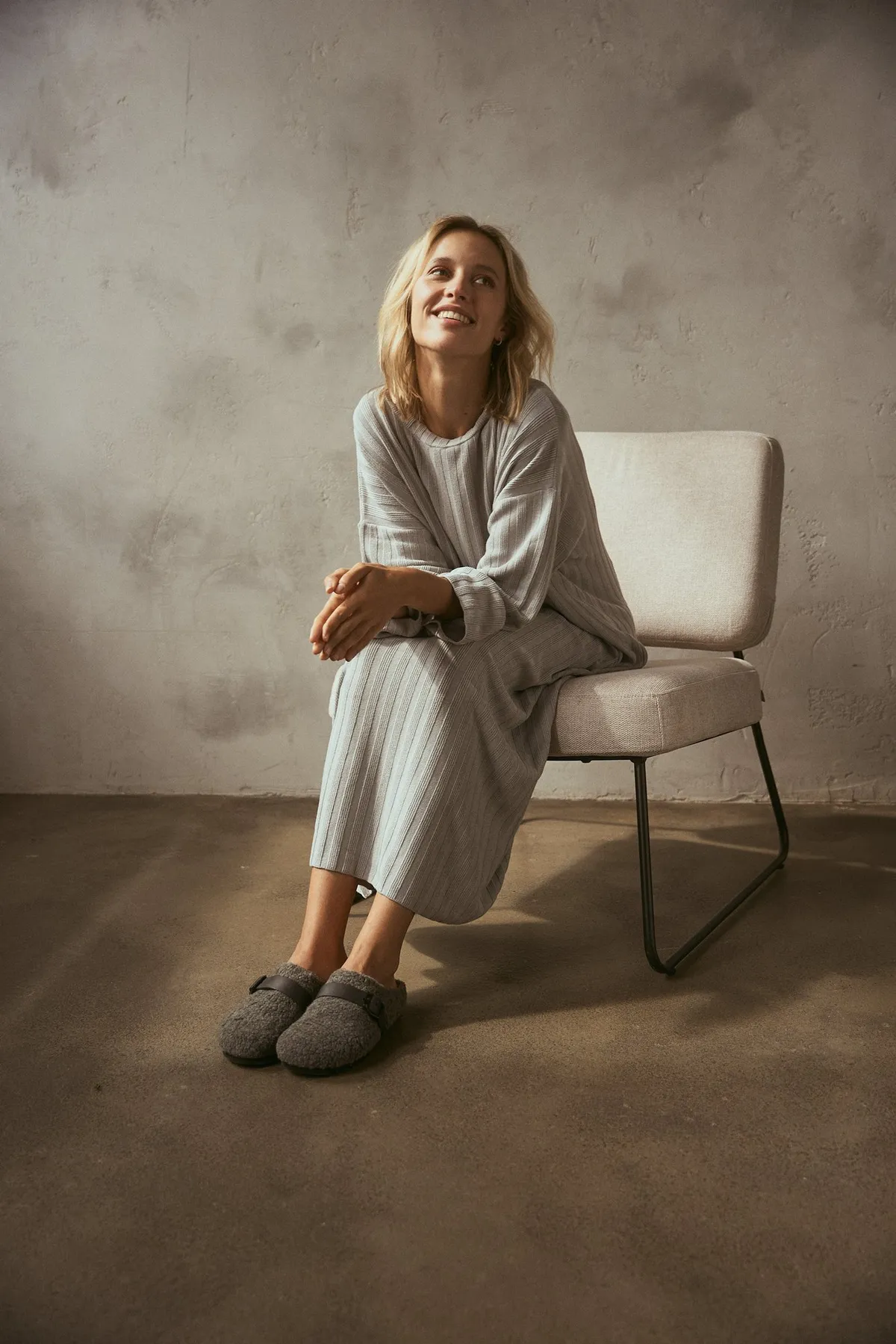 Scholl Comfort, smiling blonde model in a grey knit set with grey felt slippers on an armchair, warm wall, studio July 2022, photographed by Kilian Amrehn