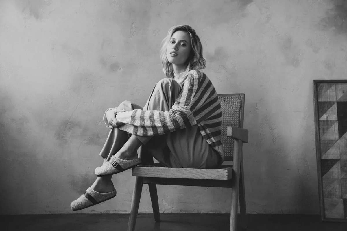 Scholl Comfort, blonde model in a striped knit sweater with fur-lined slippers on a wooden chair, black and white, studio July 2022, photographed by Kilian Amrehn