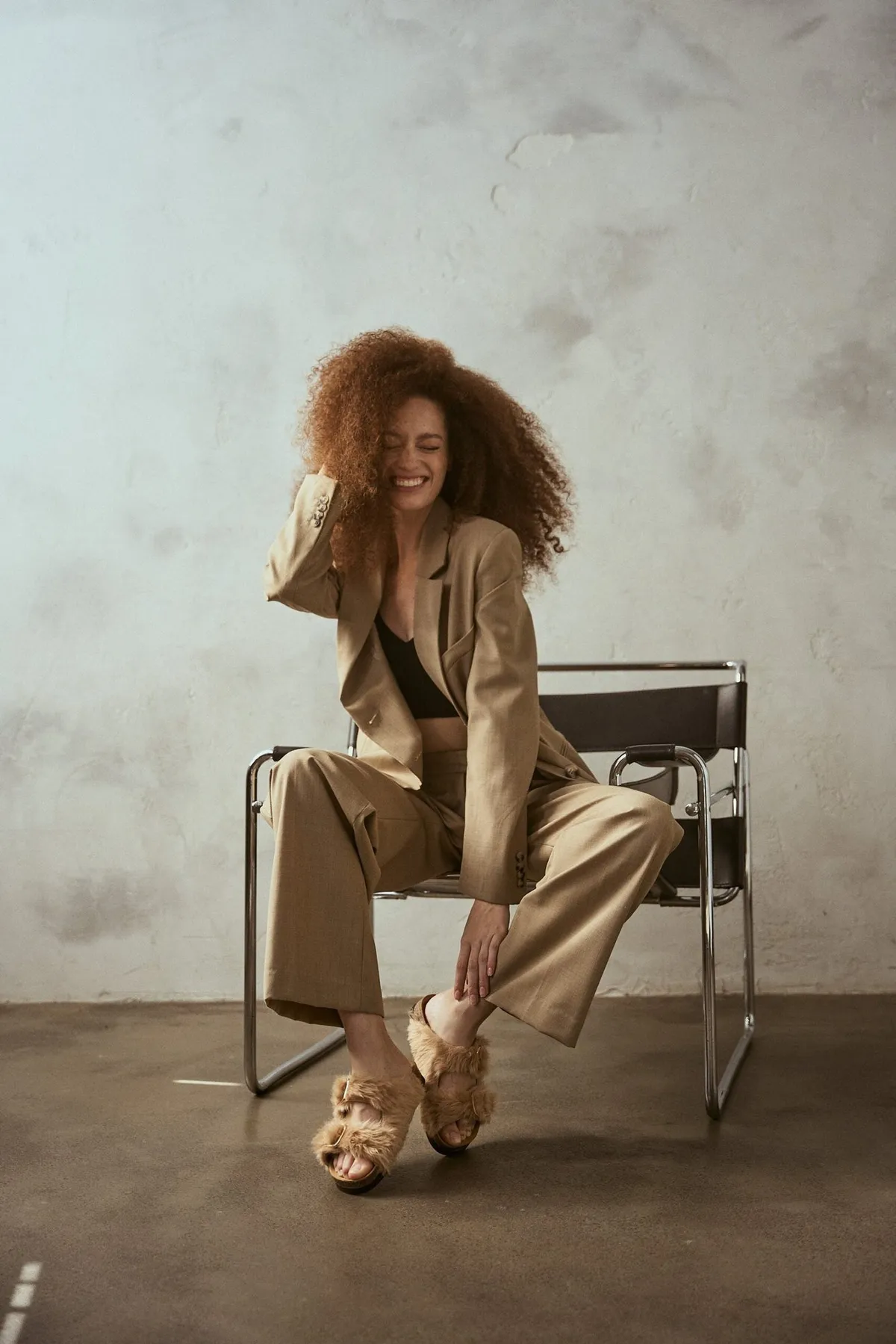 Scholl Comfort, laughing model with red curls in a beige suit and fluffy slides on a cantilever chair, studio July 2022, photographed by Kilian Amrehn