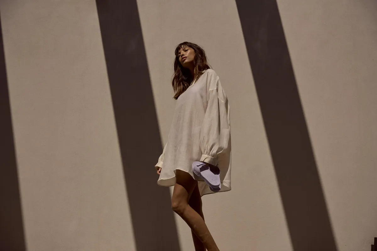Scholl Comfort, model in a loose linen shirt dress lifting a foot in a lilac slide, hard shadow stripes on the wall, Egypt 2023, photographed by Kilian Amrehn
