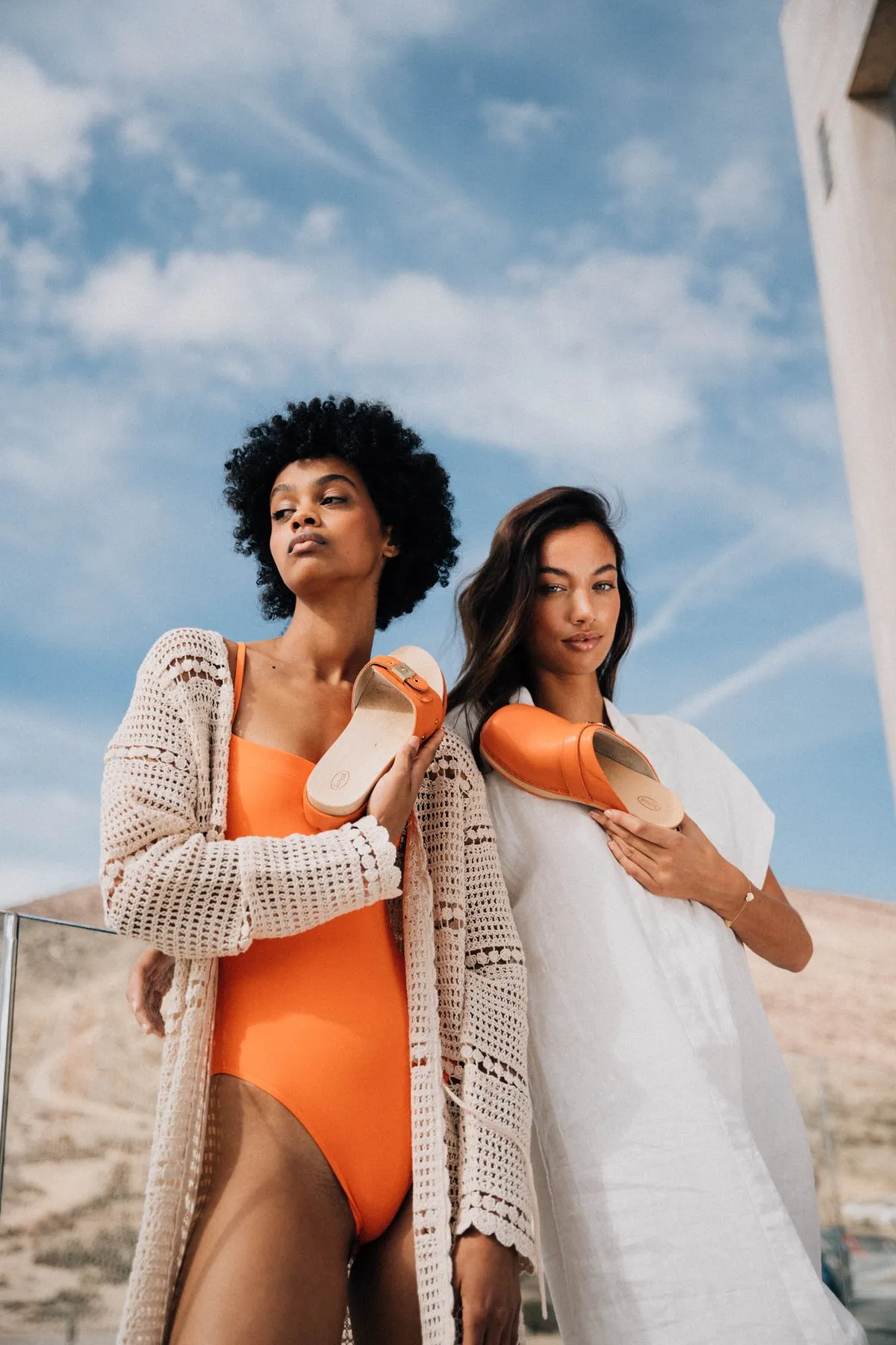 Scholl iconic, two models in swimwear and cardigans holding orange slides against blue sky, campaign Fuerteventura 2021, photographed by Kilian Amrehn