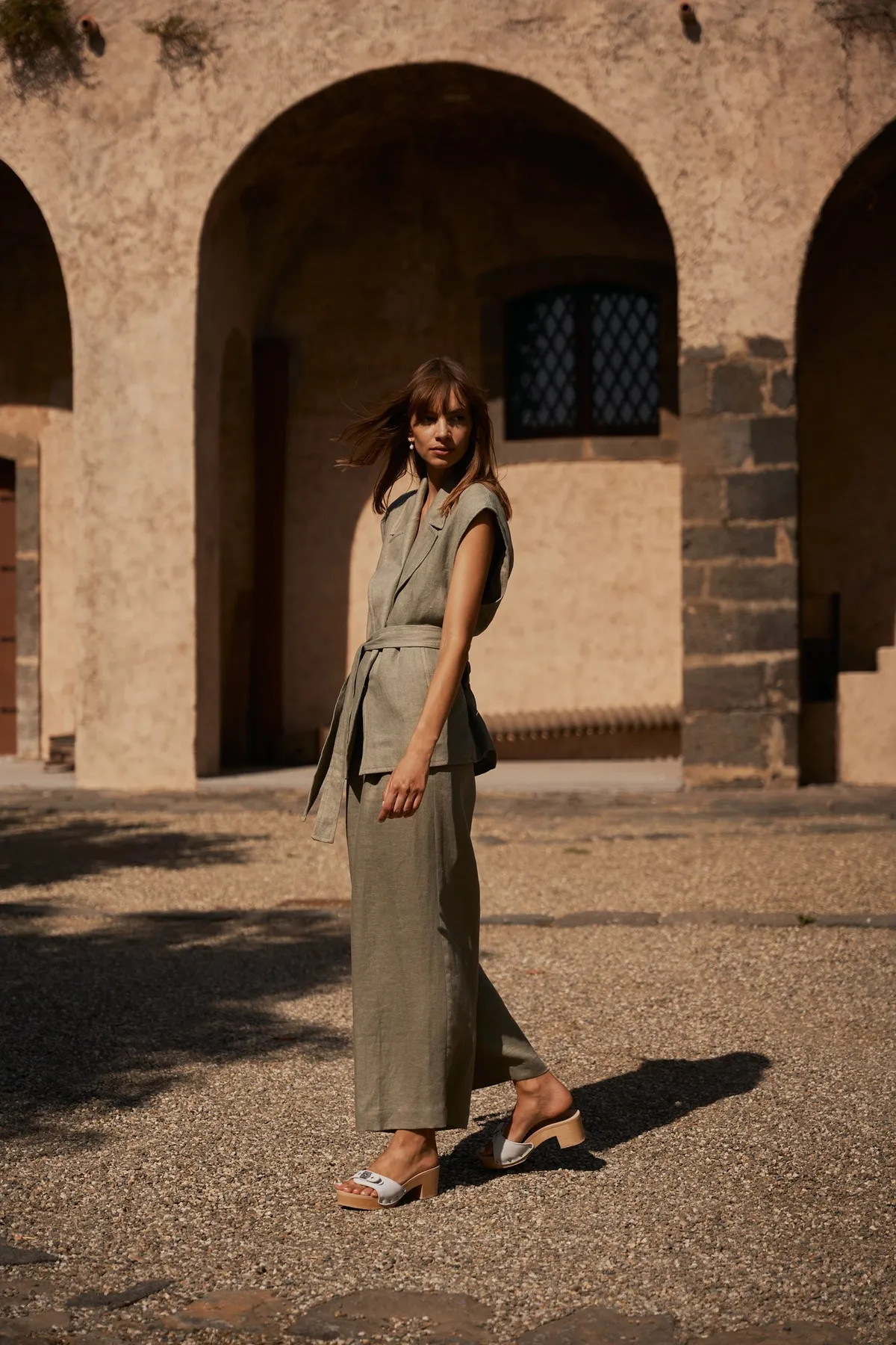 Scholl iconic, model in a khaki linen set with wooden clogs before stone arcades in warm light, campaign Sicily 2023, photographed by Kilian Amrehn