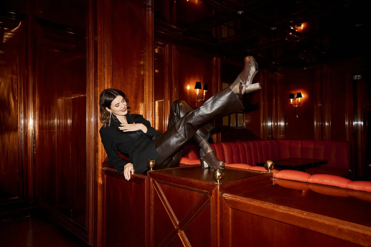 Scholl Comfort, model in a black blazer and leather trousers with heeled clogs propped on a bar counter, wood-panelled bar, Christmas campaign 2023, photographed by Kilian Amrehn