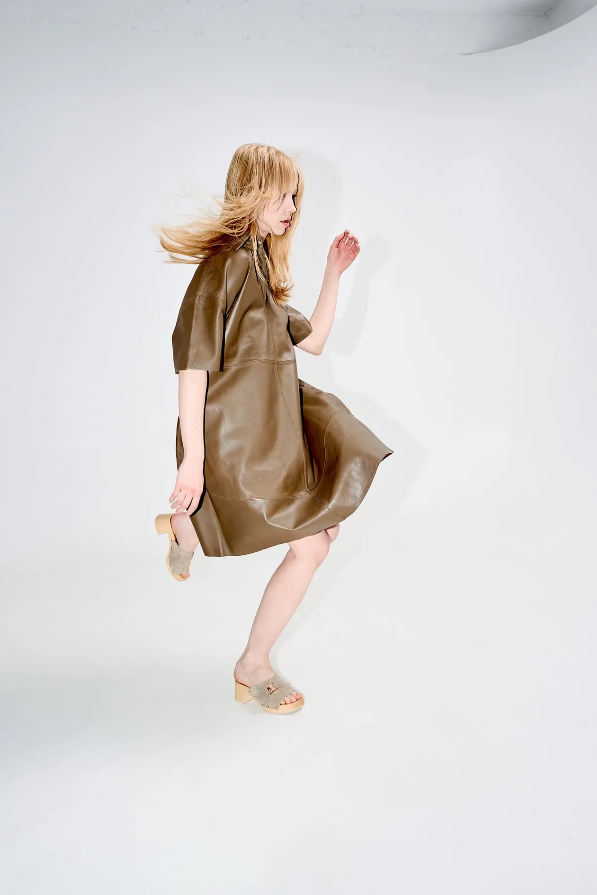 Scholl iconic, blonde model in a brown leather dress dancing in wooden clogs against a white studio backdrop, campaign February 2023, photographed by Kilian Amrehn