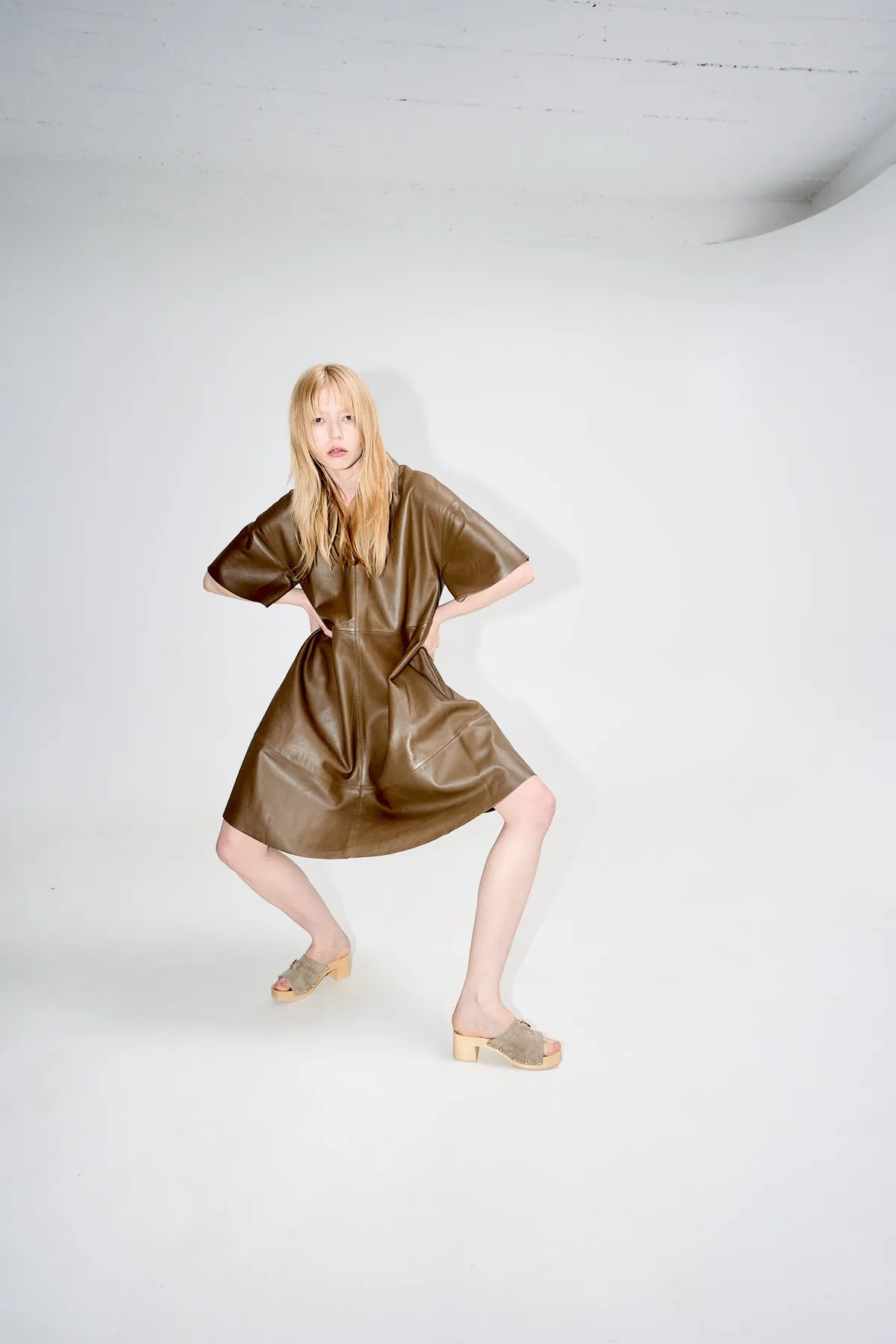 Scholl iconic, blonde model in a brown leather dress in a wide-legged stance with wooden clogs, white studio, campaign February 2023, photographed by Kilian Amrehn