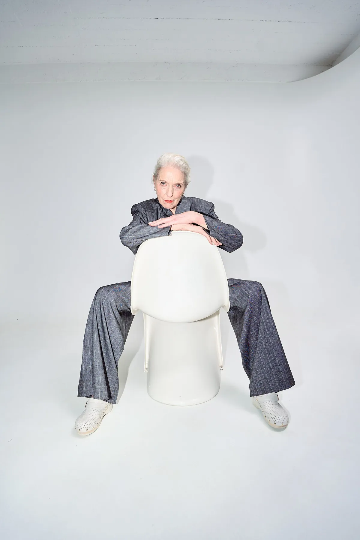 Scholl iconic, white-haired model in a grey pinstripe suit sitting backwards on a white chair, bright studio, campaign February 2023, photographed by Kilian Amrehn