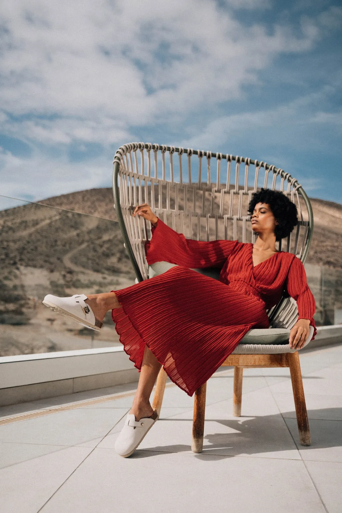 Scholl iconic, model in a red pleated dress and white clogs reclining in a rattan chair on a terrace, campaign Fuerteventura 2021, photographed by Kilian Amrehn