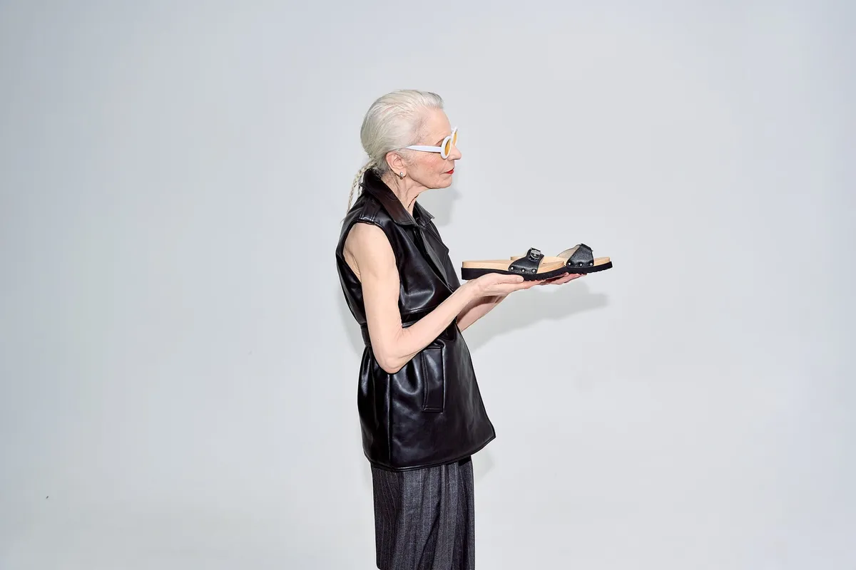 Scholl iconic, white-haired model in sunglasses and a leather vest holding a black wooden sandal on her palm, campaign February 2023, photographed by Kilian Amrehn