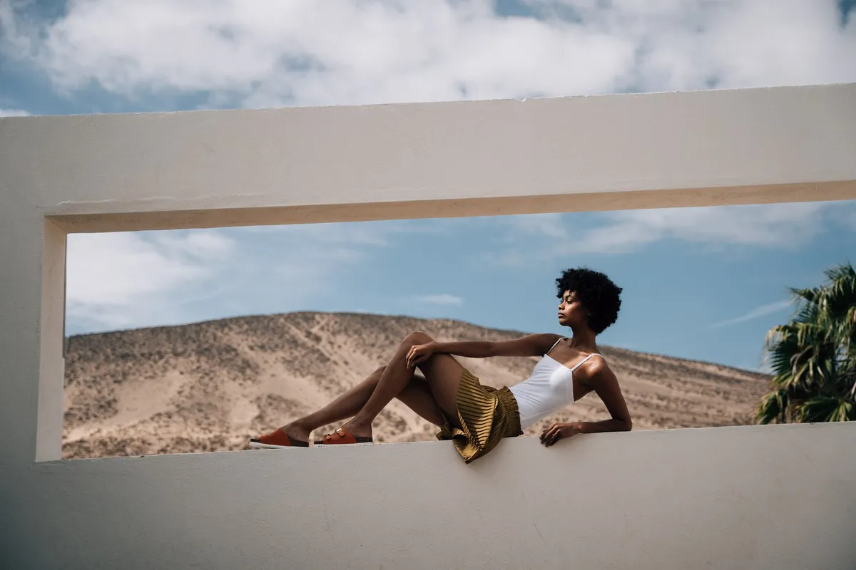 Scholl iconic, model in a white top and pleated skirt lying with sandals within a wall frame before desert hills, campaign Fuerteventura 2021, photographed by Kilian Amrehn