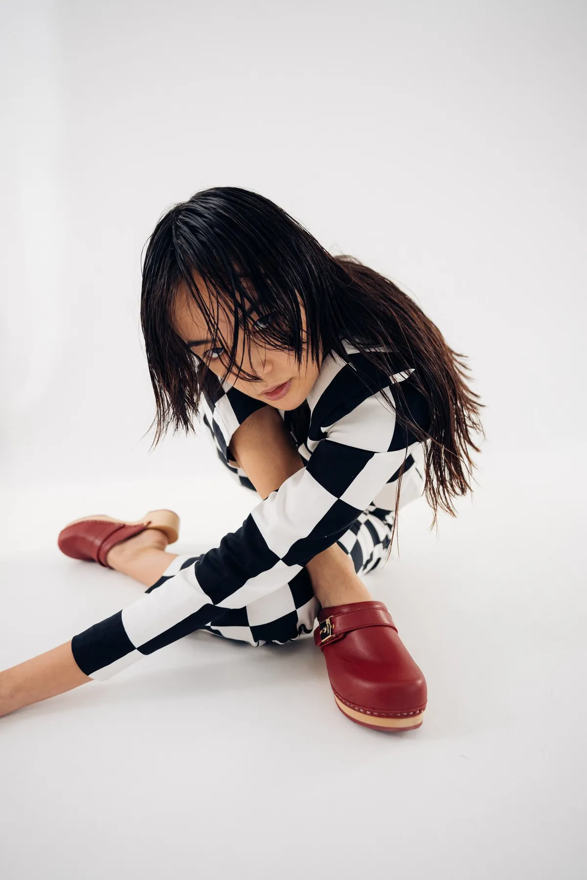 Scholl iconic, model with wet hair in a checkerboard knit sitting on the floor in red wooden clogs, light studio backdrop, campaign October 2021, photographed by Kilian Amrehn