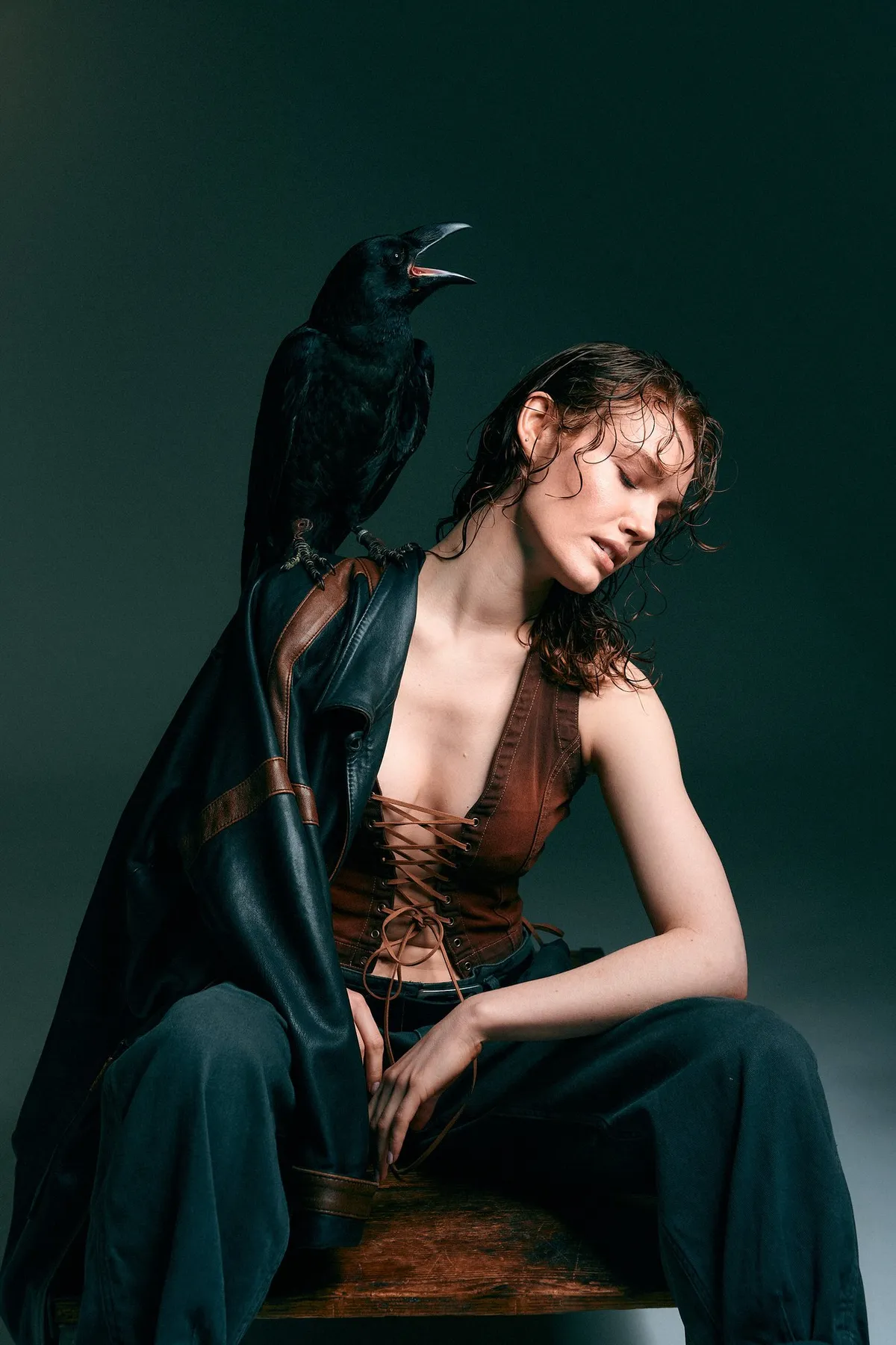 Untamed, model with wet hair in a laced leather vest and open coat, a raven on her shoulder, dark green backdrop, editorial 2024, photographed by Kilian Amrehn