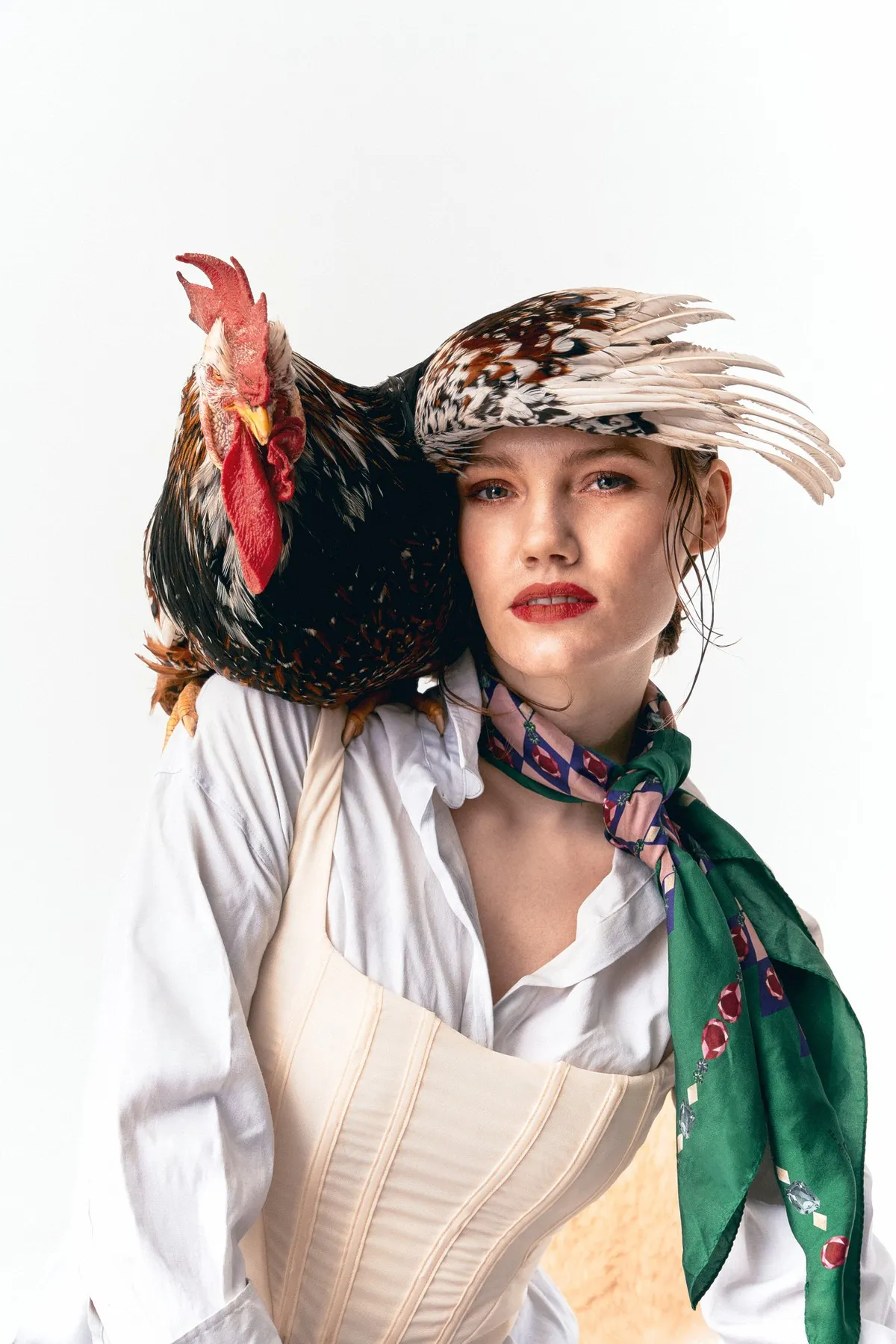 Untamed, model in a feathered cap, corset and green silk scarf with a rooster on her shoulder, red lipstick against white, editorial 2024, photographed by Kilian Amrehn