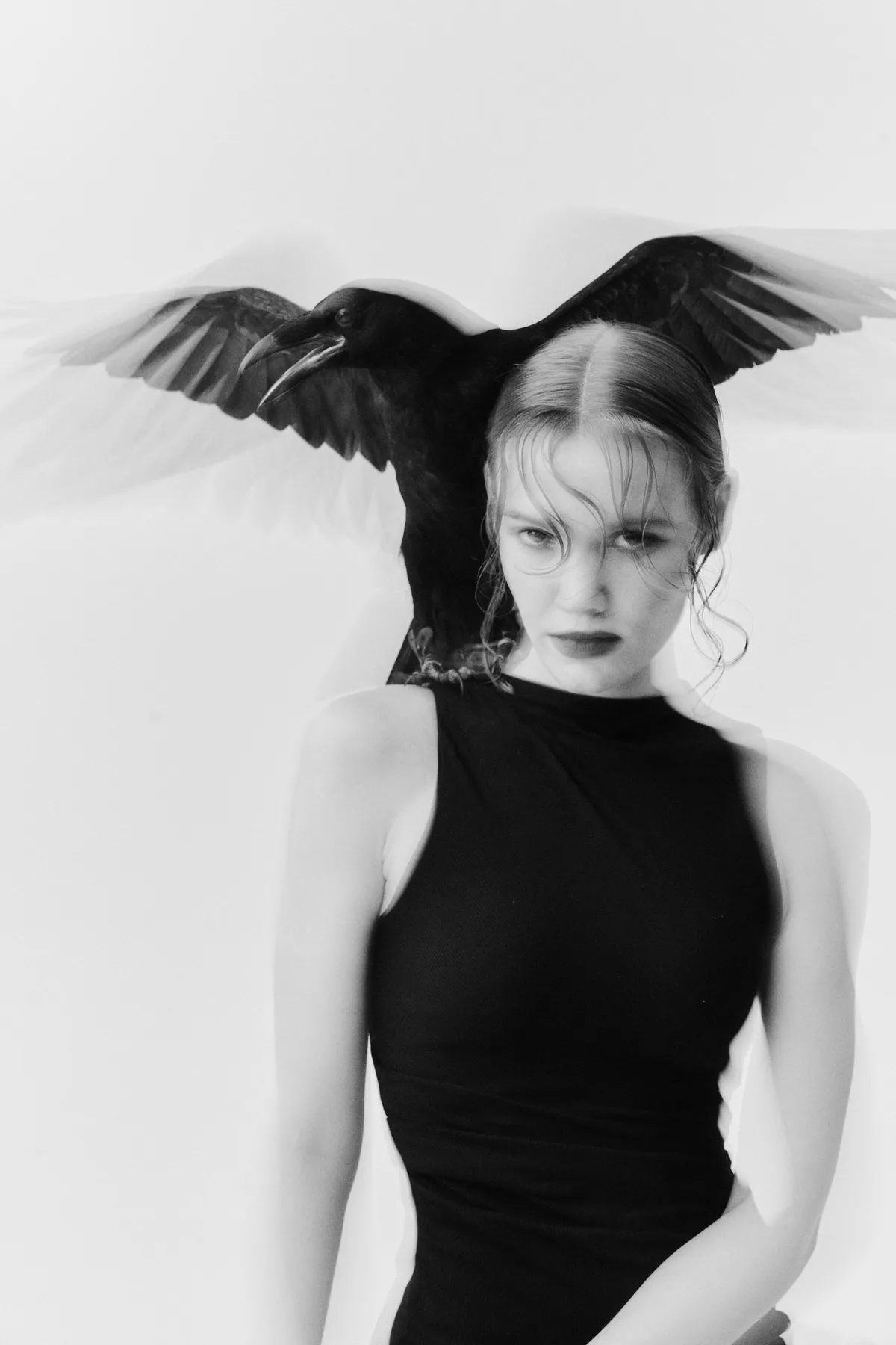 Untamed, raven spreading its wings above the head of a model in a black bodysuit, serious gaze, black and white, editorial 2024, photographed by Kilian Amrehn
