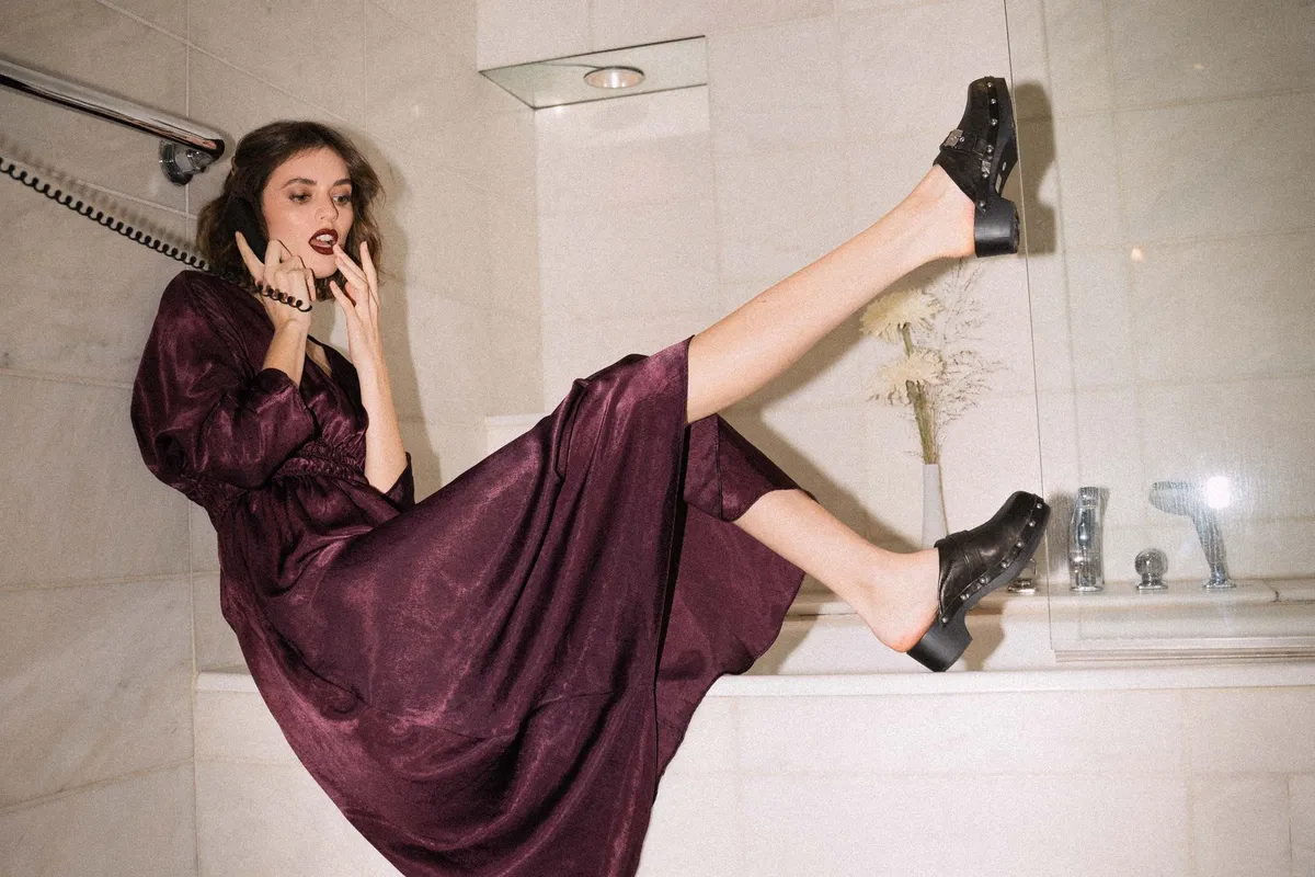 The Wants, model in a burgundy satin dress on the phone in a bathtub, legs up in black studded clogs, campaign 2020, photographed by Kilian Amrehn
